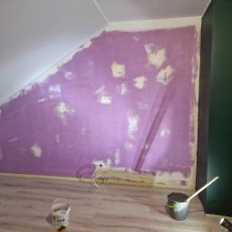 BAJ DEVELOPMENT & CREATIVE SPÓŁKA Z OGRANICZONĄ ODPOWIEDZIALNOŚCIĄ - Ściana w pokoju na poddaszu po szpachlowaniu, przygotowana do malowania, z widocznymi plamami szpachli i niedomalowaniami, zabezpieczona taśmą malarską przy podłodze.