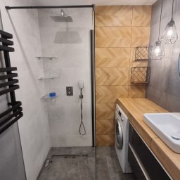 BAJ DEVELOPMENT & CREATIVE SPÓŁKA Z OGRANICZONĄ ODPOWIEDZIALNOŚCIĄ - Nowoczesna łazienka z kabiną prysznicową typu walk-in z czarną ramą, grzejnikiem drabinkowym, drewnopodobnymi płytkami ułożonymi w jodełkę na ścianie, pralką pod blatem i wiszącymi lampami...