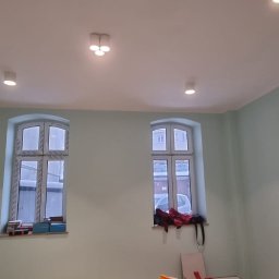 BAJ DEVELOPMENT & CREATIVE SPÓŁKA Z OGRANICZONĄ ODPOWIEDZIALNOŚCIĄ - Świeżo pomalowane wnętrze pokoju z dwoma oknami, ściany w odcieniu pastelowej zieleni, biały sufit z trzema punktami oświetleniowymi.
