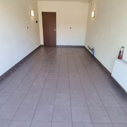 BAJ DEVELOPMENT & CREATIVE SPÓŁKA Z OGRANICZONĄ ODPOWIEDZIALNOŚCIĄ - Wykafelkowana podłoga garażu z brązowych płytek, widoczny fragment grzejnika i instalacji wodnej przy ścianie, brązowe listwy przypodłogowe.