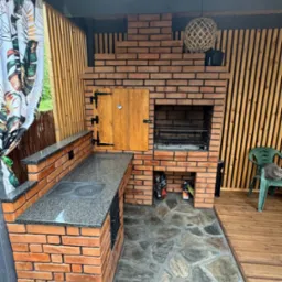 Murowany grill z cegły z blatem z szarego granitu, drewnianymi drzwiczkami i kominem, obok drewniana ściana z pionowych desek i kot siedzący na zielonym krześle.