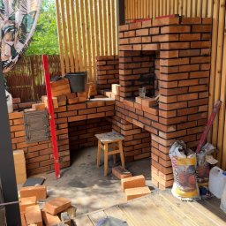 BAJ DEVELOPMENT & CREATIVE SPÓŁKA Z OGRANICZONĄ ODPOWIEDZIALNOŚCIĄ - Budowa murowanego grilla z cegły na zewnątrz, widoczny poziomica, wiadro, zaprawa, narzędzia i drewniane wykończenie altany.