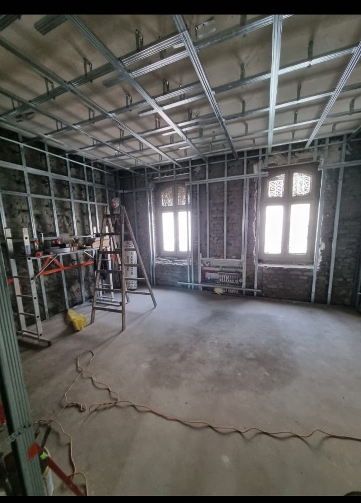 Wnętrze w trakcie prac wykończeniowych: surowy beton na podłodze, ściany z odsłoniętą cegłą i metalową konstrukcją pod zabudowę, dwie okiennice z dekoracyjnymi wzorami, drabina i narzędzia budowlane.