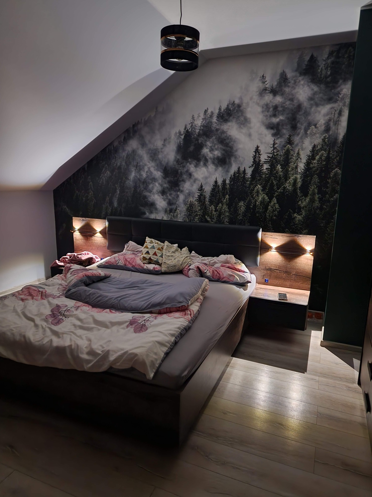 Sypialnia na poddaszu z fototapetą przedstawiającą mglisty las, drewnianymi szafkami nocnymi z oświetleniem punktowym i łóżkiem z szarą pościelą w kwiaty.