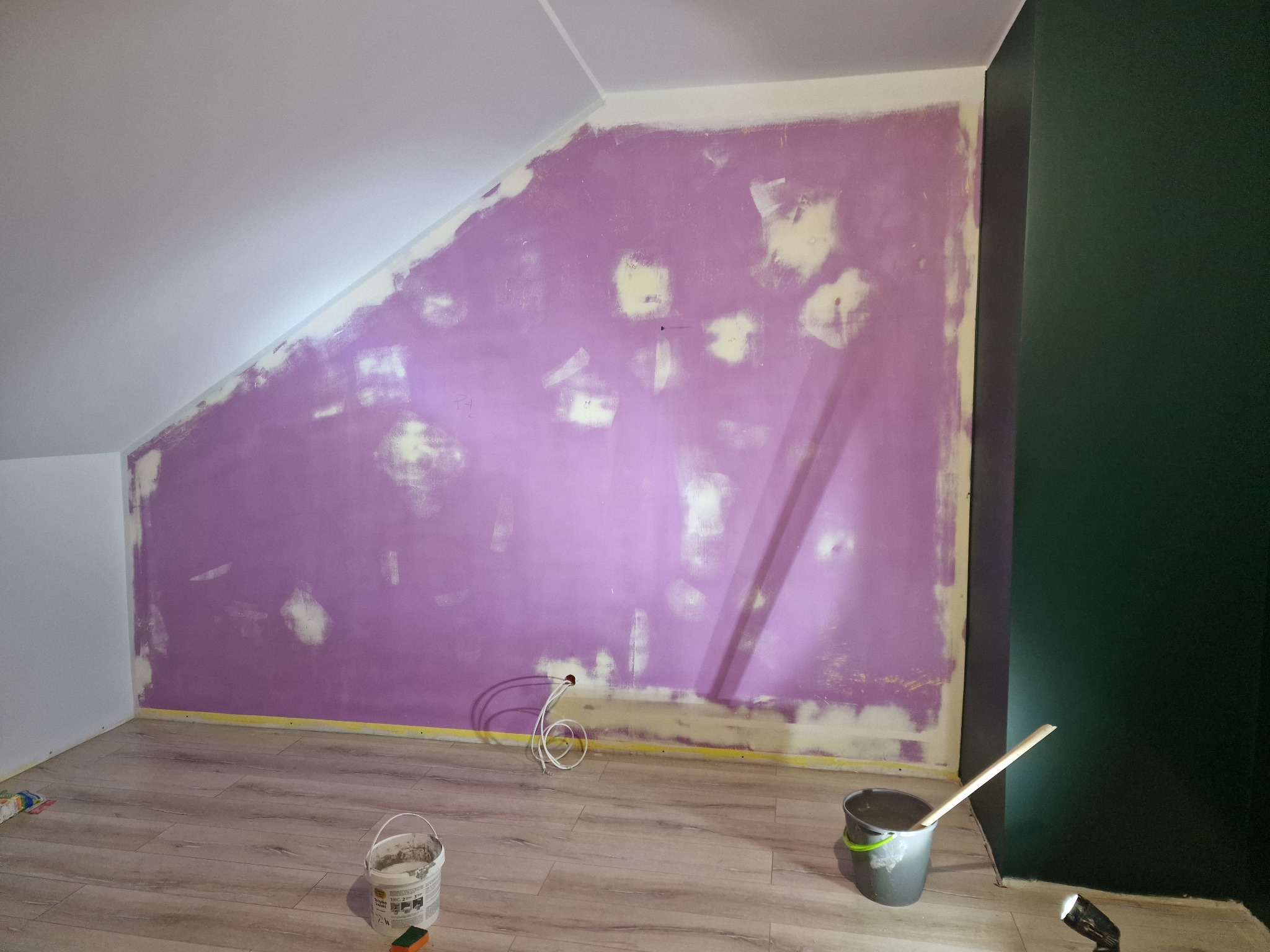 Ściana w pokoju na poddaszu po szpachlowaniu, przygotowana do malowania, z widocznymi plamami szpachli i niedomalowaniami, zabezpieczona taśmą malarską przy podłodze.