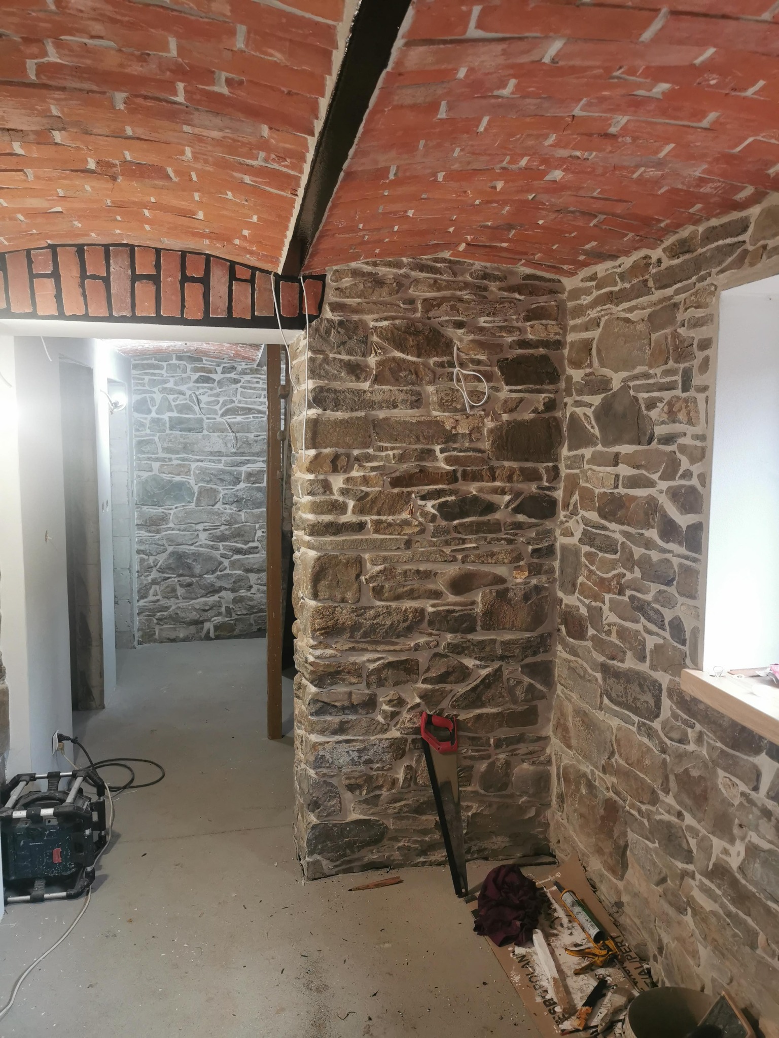 Piwnica z surowymi ścianami z kamienia, ceglany łuk sklepienia i narzędzia budowlane na podłodze, w tym piła ręczna i kuweta z zaprawą.