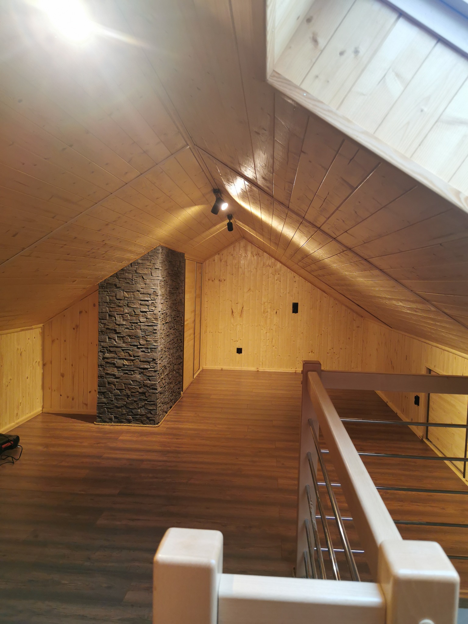 Wykończone poddasze ze ścianami i sufitem pokrytymi drewnianą boazerią, podłoga z paneli, komin obłożony kamieniem dekoracyjnym, oświetlenie punktowe i drewniana balustrada.