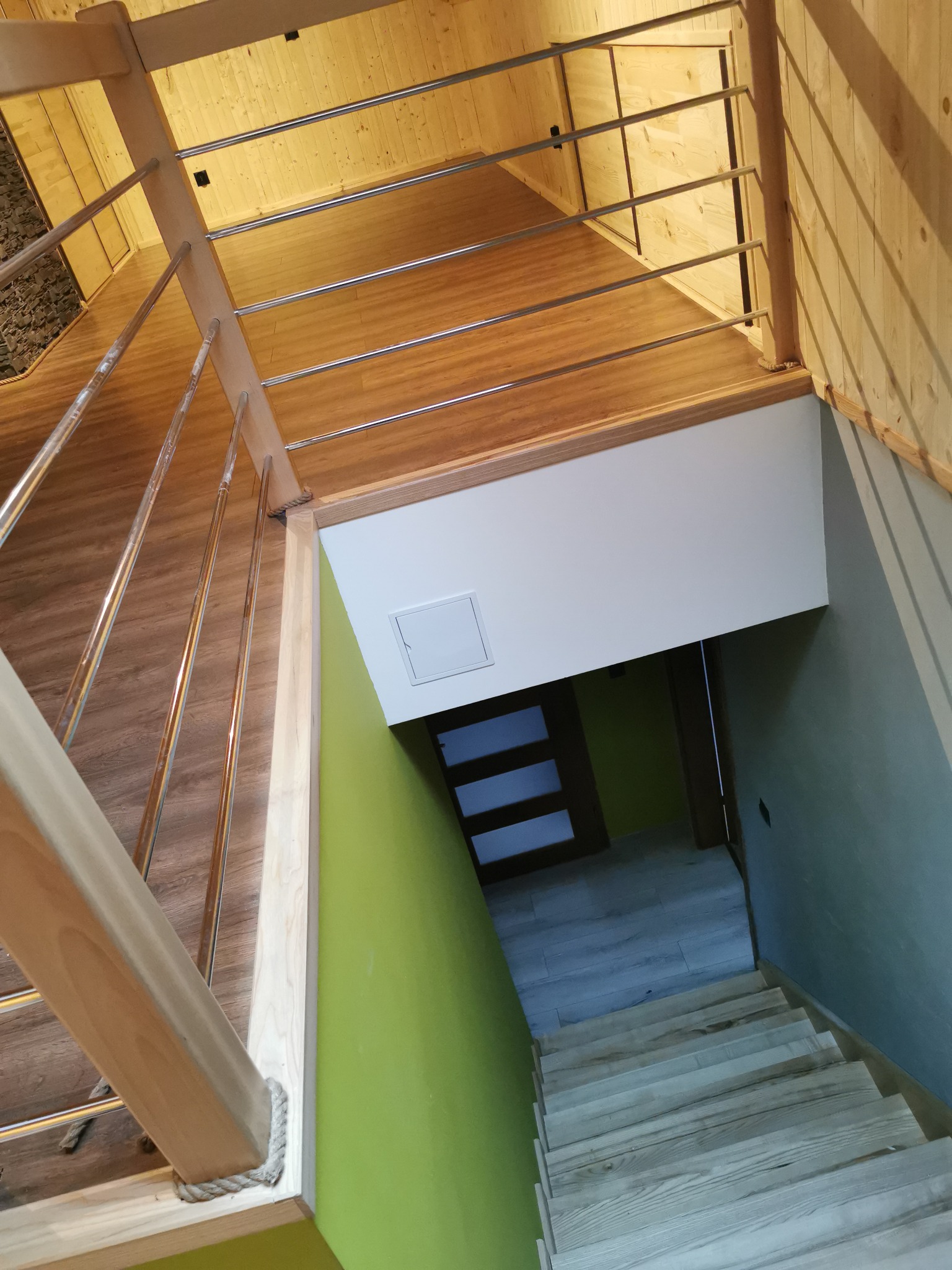 Wnętrze domu w trakcie wykańczania, widok z góry na drewniane schody z metalową balustradą, zielona ściana, drewniana boazeria na suficie i fragment podłogi z panelami.