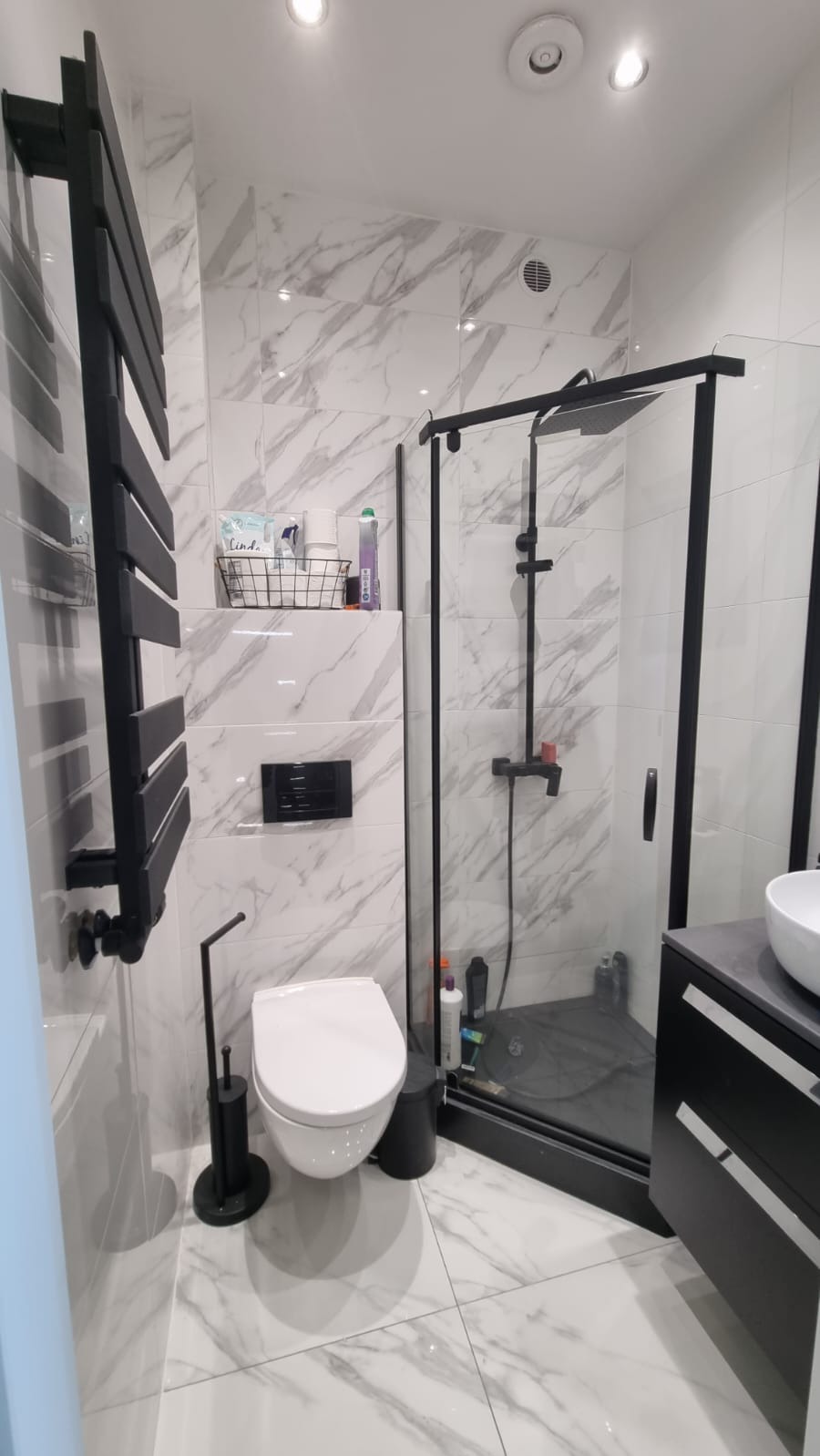 Nowoczesna łazienka z białymi płytkami imitującymi marmur, czarną kabiną prysznicową i grzejnikiem drabinkowym w tym samym kolorze, wisząca toaleta i szafka z umywalką.