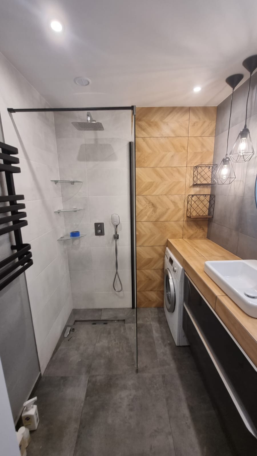 Nowoczesna łazienka z kabiną prysznicową typu walk-in z czarną ramą, grzejnikiem drabinkowym, drewnopodobnymi płytkami ułożonymi w jodełkę na ścianie, pralką pod blatem i wiszącymi lampami...