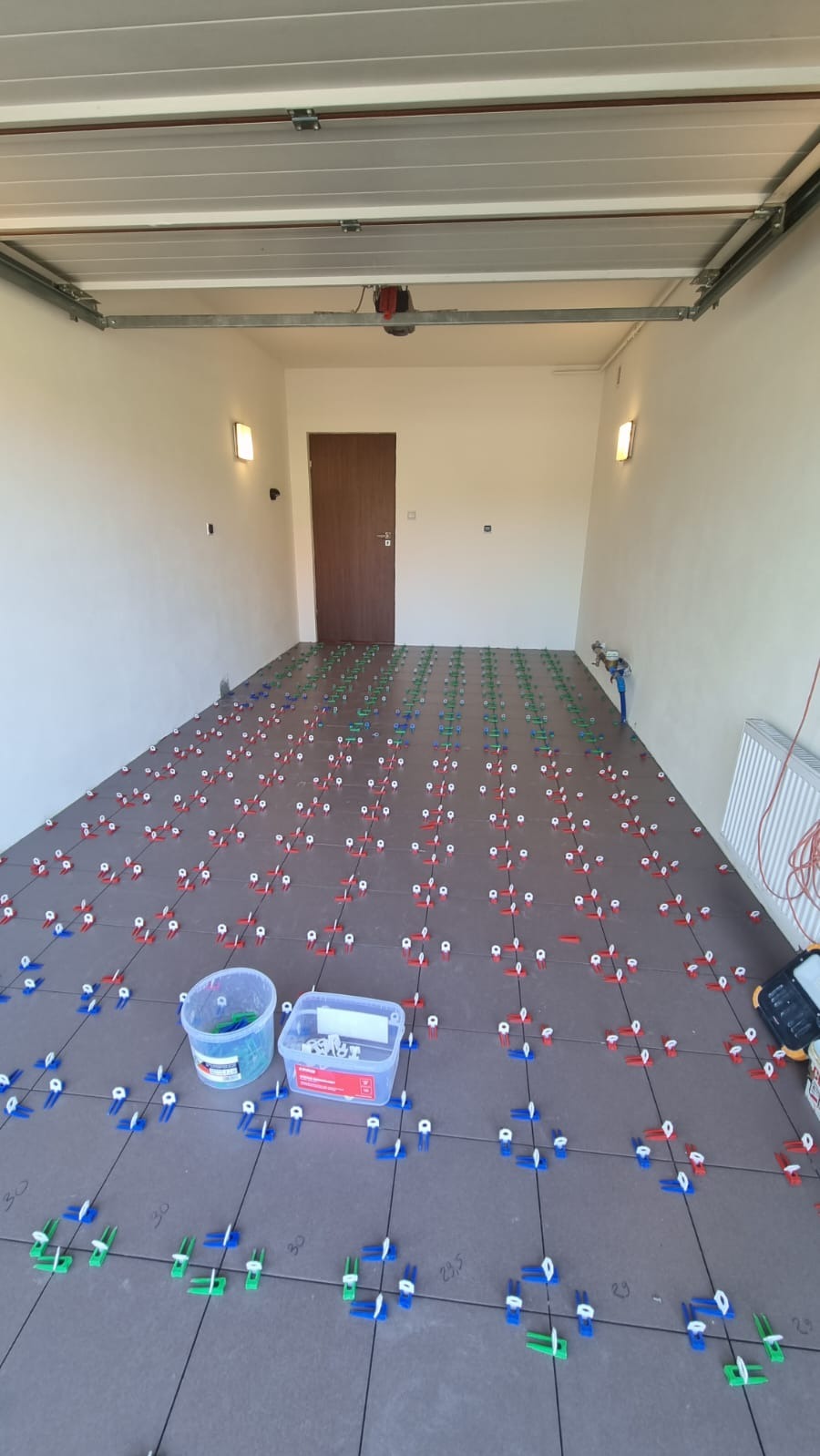 Układanie ciemnoszarych płytek podłogowych w garażu z użyciem systemu poziomowania płytek z klinami i klipsami w kolorach niebieskim, zielonym i czerwonym. Widoczne wiaderka z klipsami i pudełko...