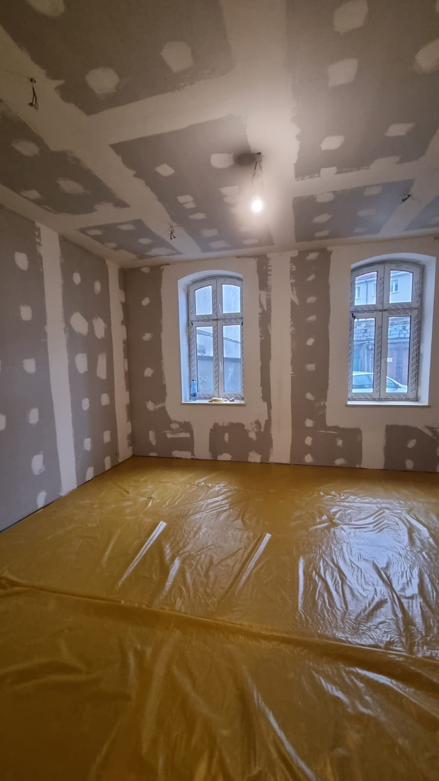Pokój w trakcie remontu: ściany z płyt gipsowo-kartonowych z widocznymi łączeniami szpachlowanymi, podłoga zabezpieczona folią ochronną, dwa okna z białymi ramami.