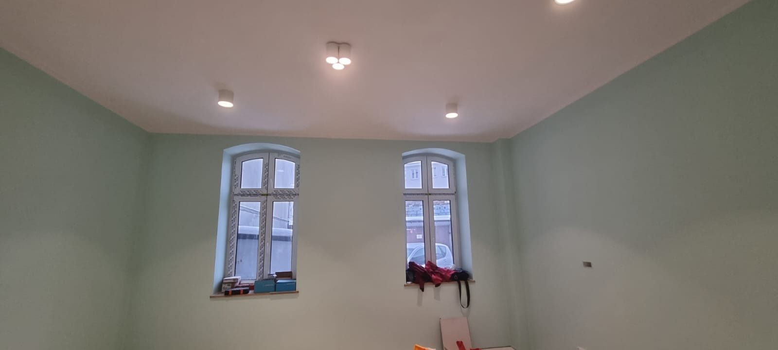 Świeżo pomalowane wnętrze pokoju z dwoma oknami, ściany w odcieniu pastelowej zieleni, biały sufit z trzema punktami oświetleniowymi.