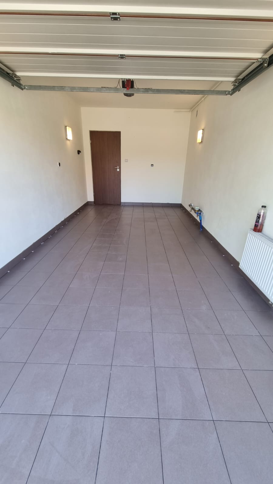 Wykafelkowana podłoga garażu z brązowych płytek, widoczny fragment grzejnika i instalacji wodnej przy ścianie, brązowe listwy przypodłogowe.