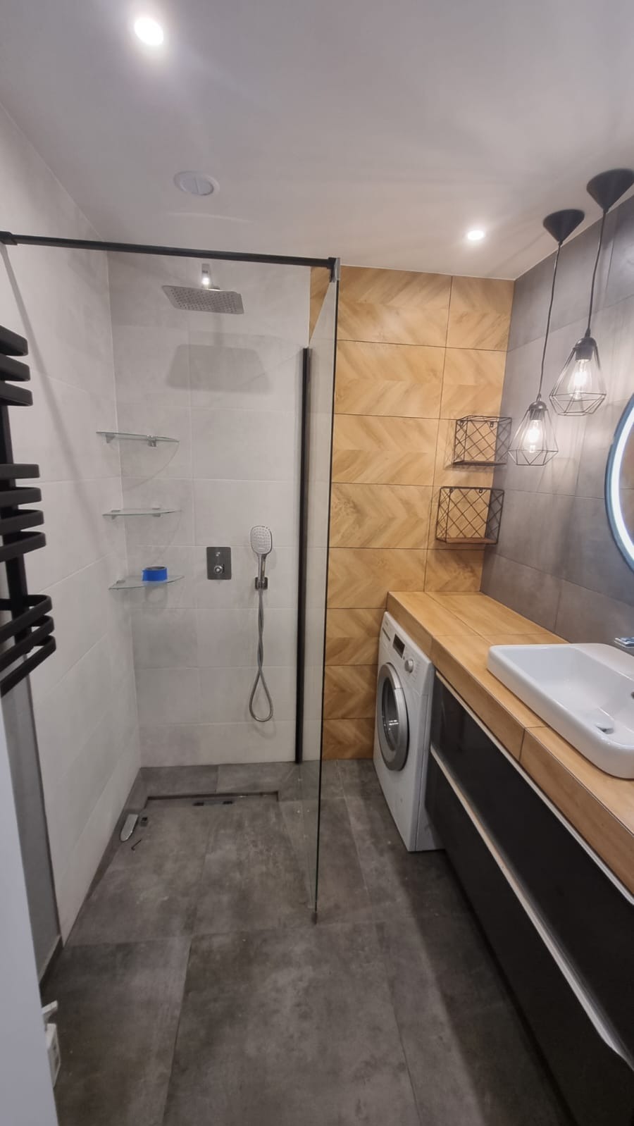 Nowoczesna łazienka z kabiną prysznicową typu walk-in, szarą płytką podłogową, drewnopodobną ścianą w jodełkę, pralką pod blatem i wiszącymi lampami drucianymi.