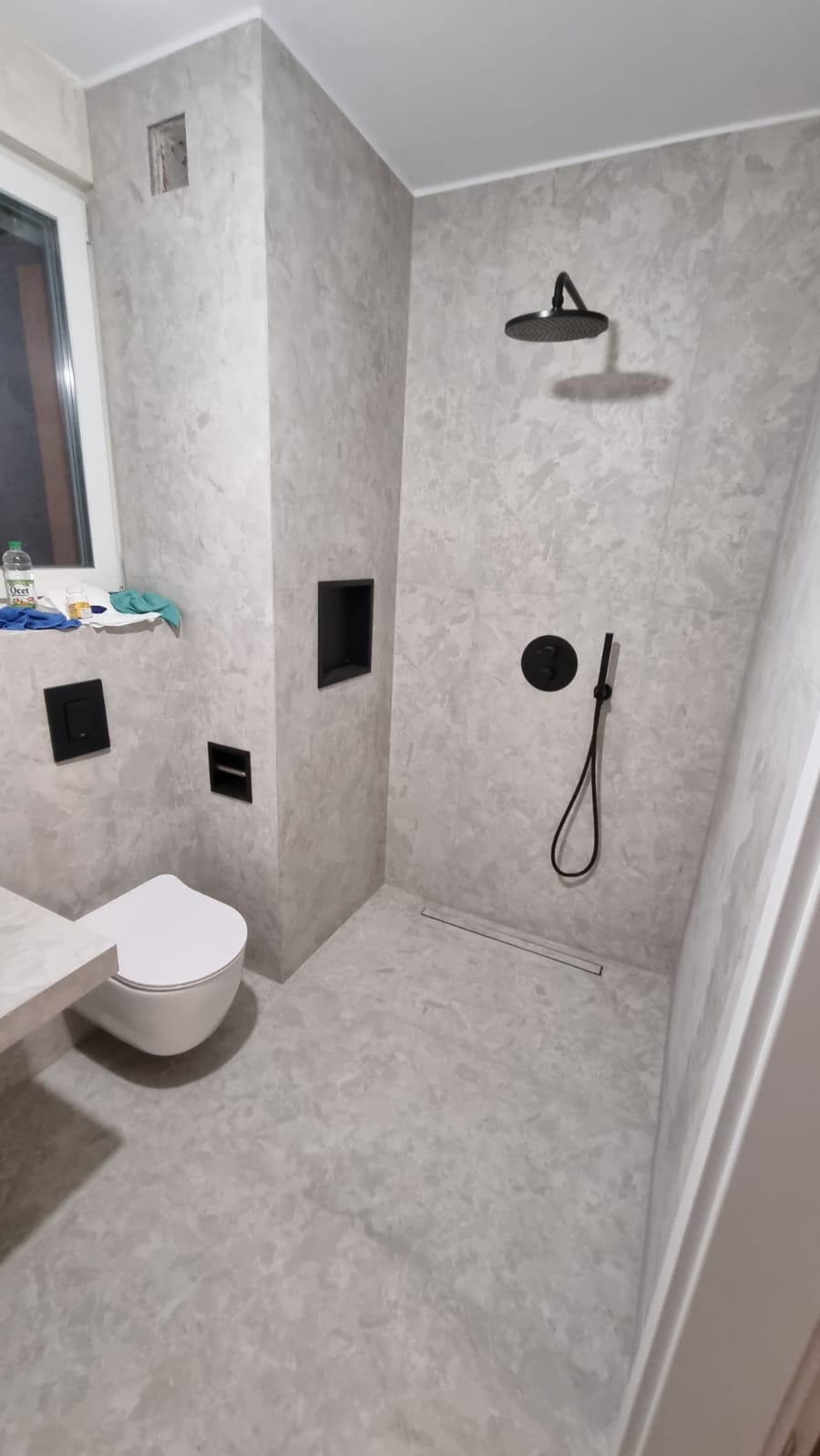 Nowoczesna łazienka z szarymi ścianami o fakturze betonu, czarną armaturą prysznicową i toaletą podwieszaną. W rogu okno z widocznymi środkami czystości.