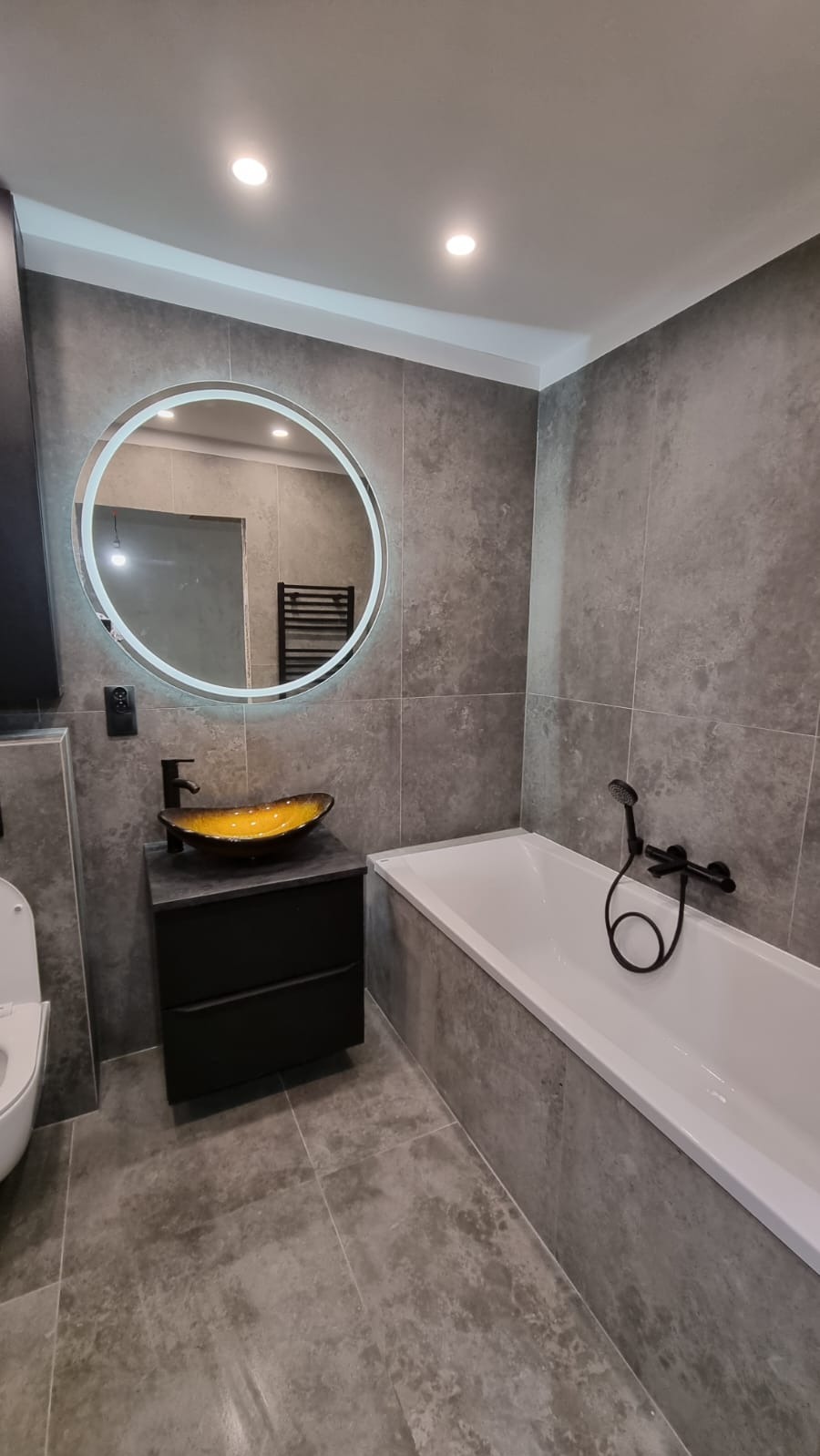 Nowoczesna łazienka z szarymi płytkami imitującymi kamień, okrągłym podświetlanym lustrem, czarną armaturą i umywalką w kolorze bursztynowym.