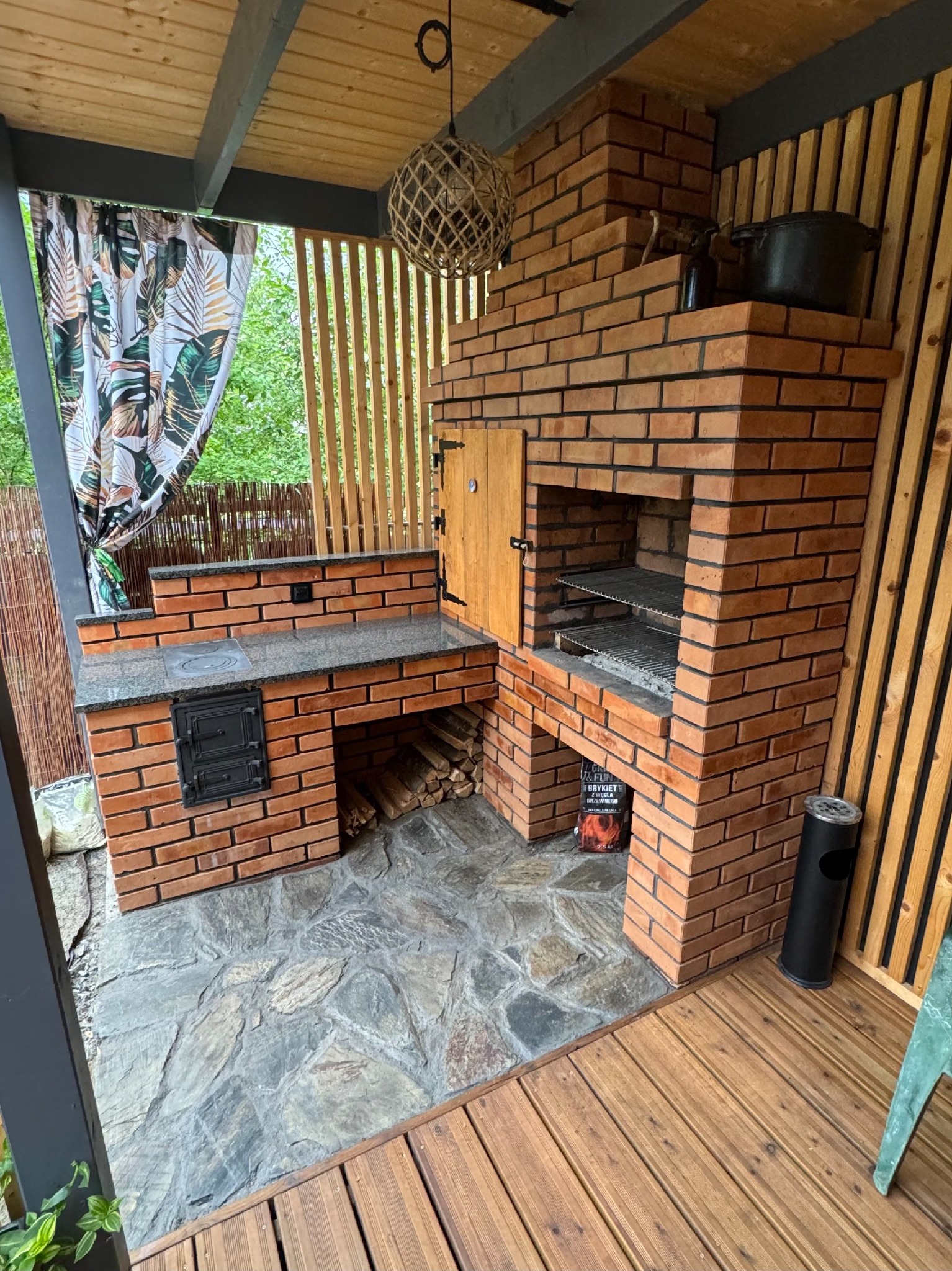 Zewnętrzna kuchnia z cegły z grillem, płytą grzejną i miejscem do przechowywania drewna, wykończona drewnianą boazerią i kamienną podłogą, oświetlona wiszącą lampą z naturalnych materiałów.