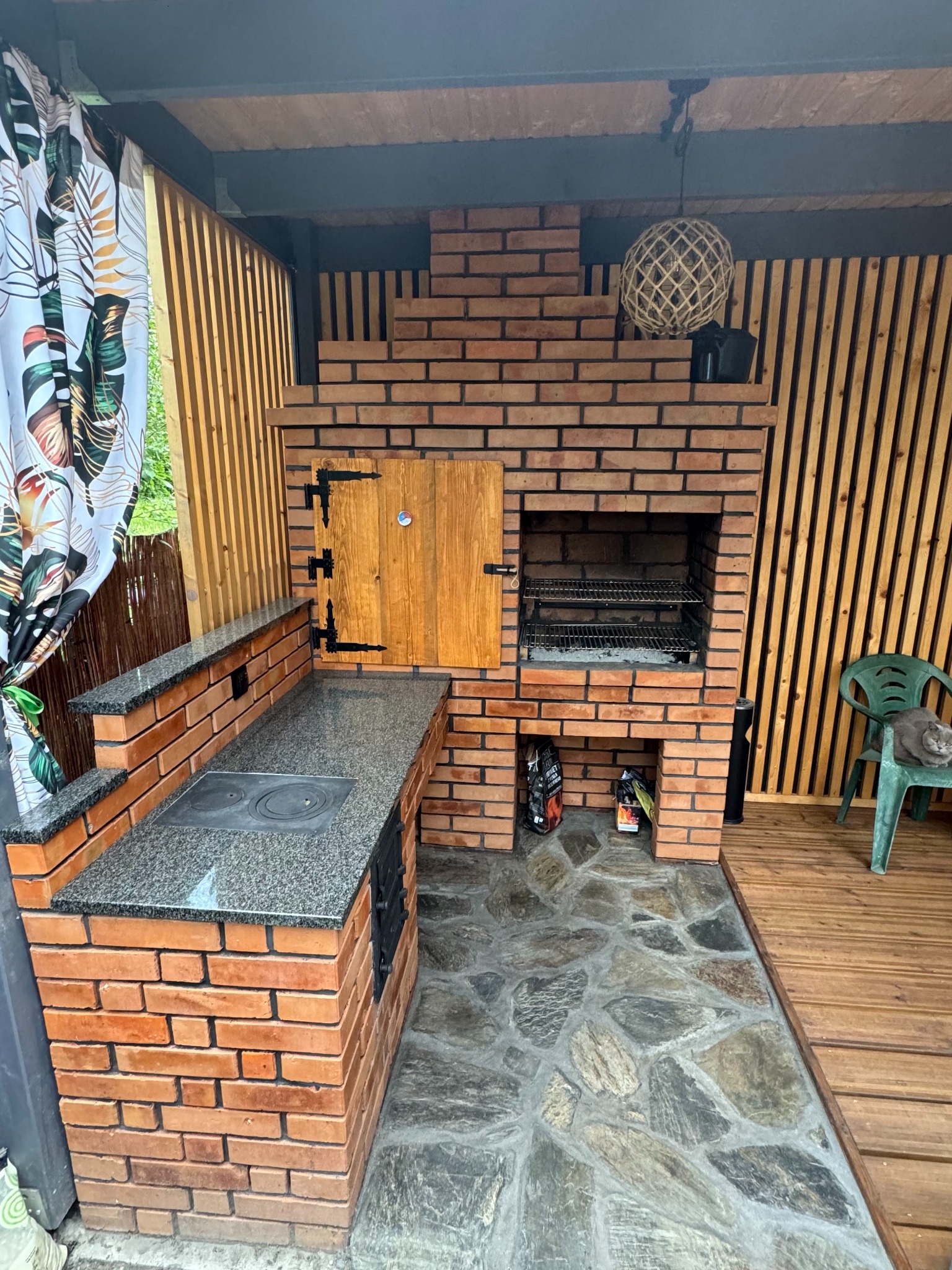 Murowany grill z cegły z blatem z szarego granitu, drewnianymi drzwiczkami i kominem, obok drewniana ściana z pionowych desek i kot siedzący na zielonym krześle.