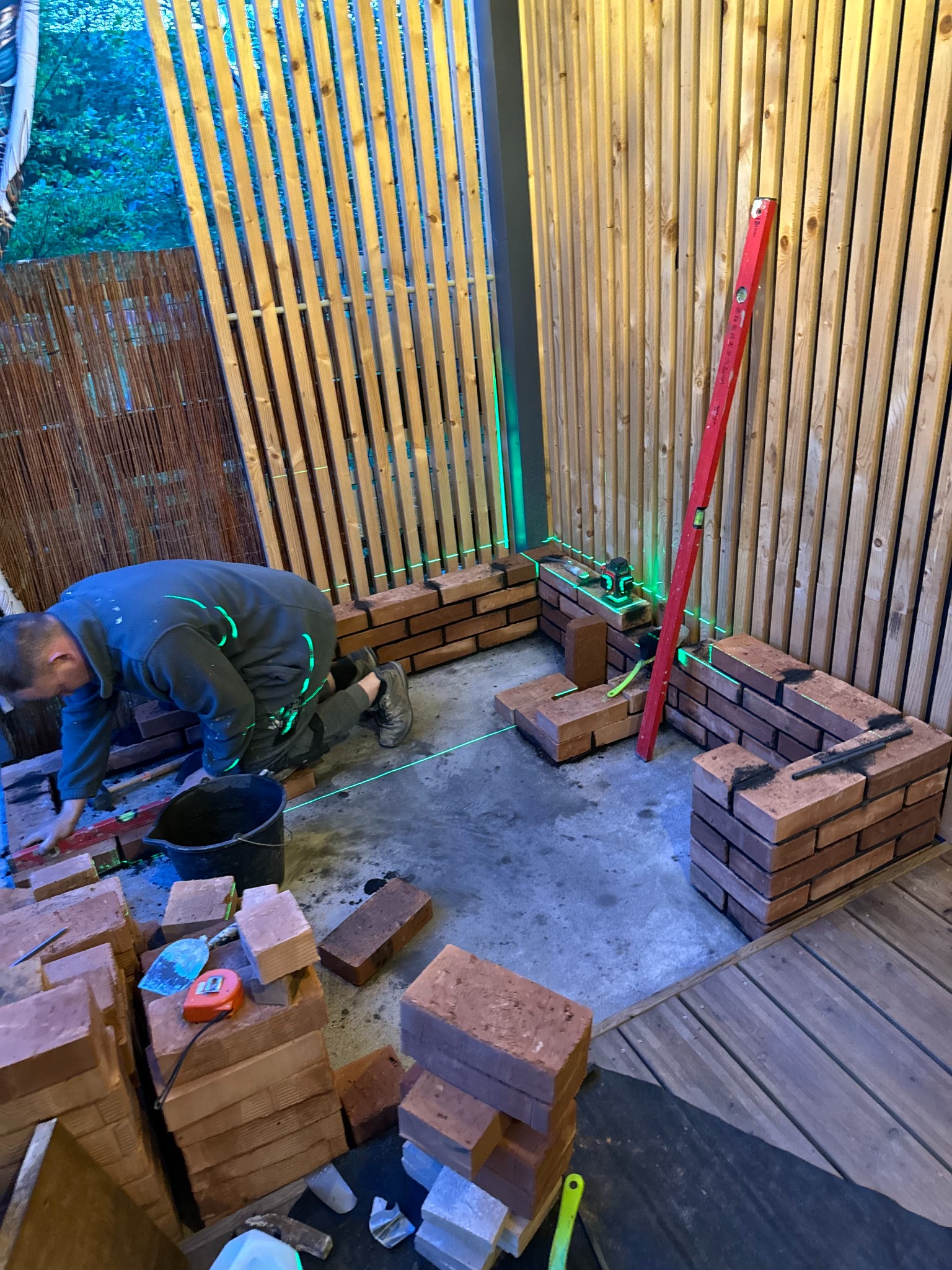 Murarz układa cegły z użyciem poziomicy laserowej w pomieszczeniu z drewnianymi ścianami, widoczny stos cegieł, poziomica i wiadro.