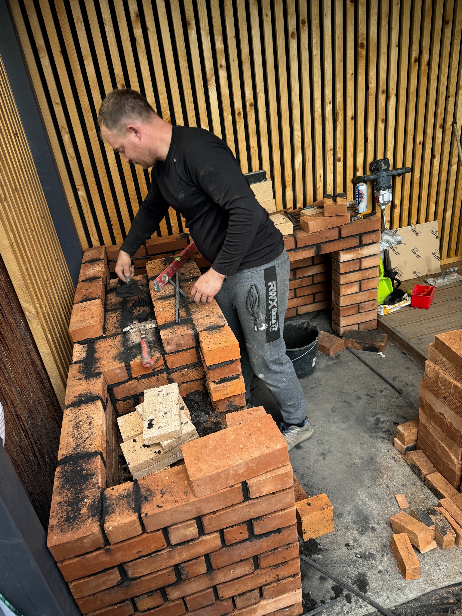 Mężczyzna w szarych dresach buduje z cegieł konstrukcję, prawdopodobnie grilla lub wędzarnię, używając kielni i zaprawy, widoczne narzędzia i materiały budowlane.