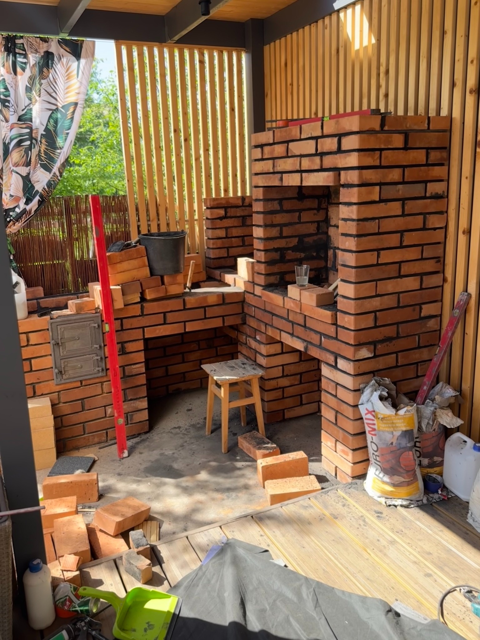 Budowa murowanego grilla z cegły na zewnątrz, widoczny poziomica, wiadro, zaprawa, narzędzia i drewniane wykończenie altany.