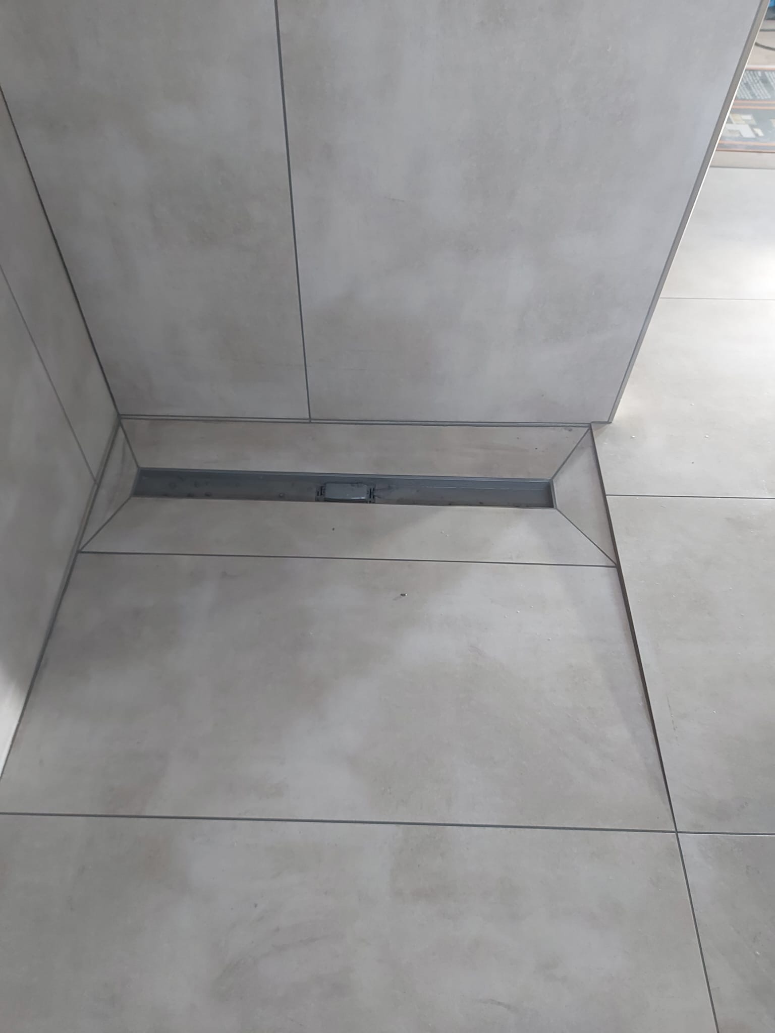 Szare, matowe płytki podłogowe i ścienne z odpływem liniowym w rogu łazienki.