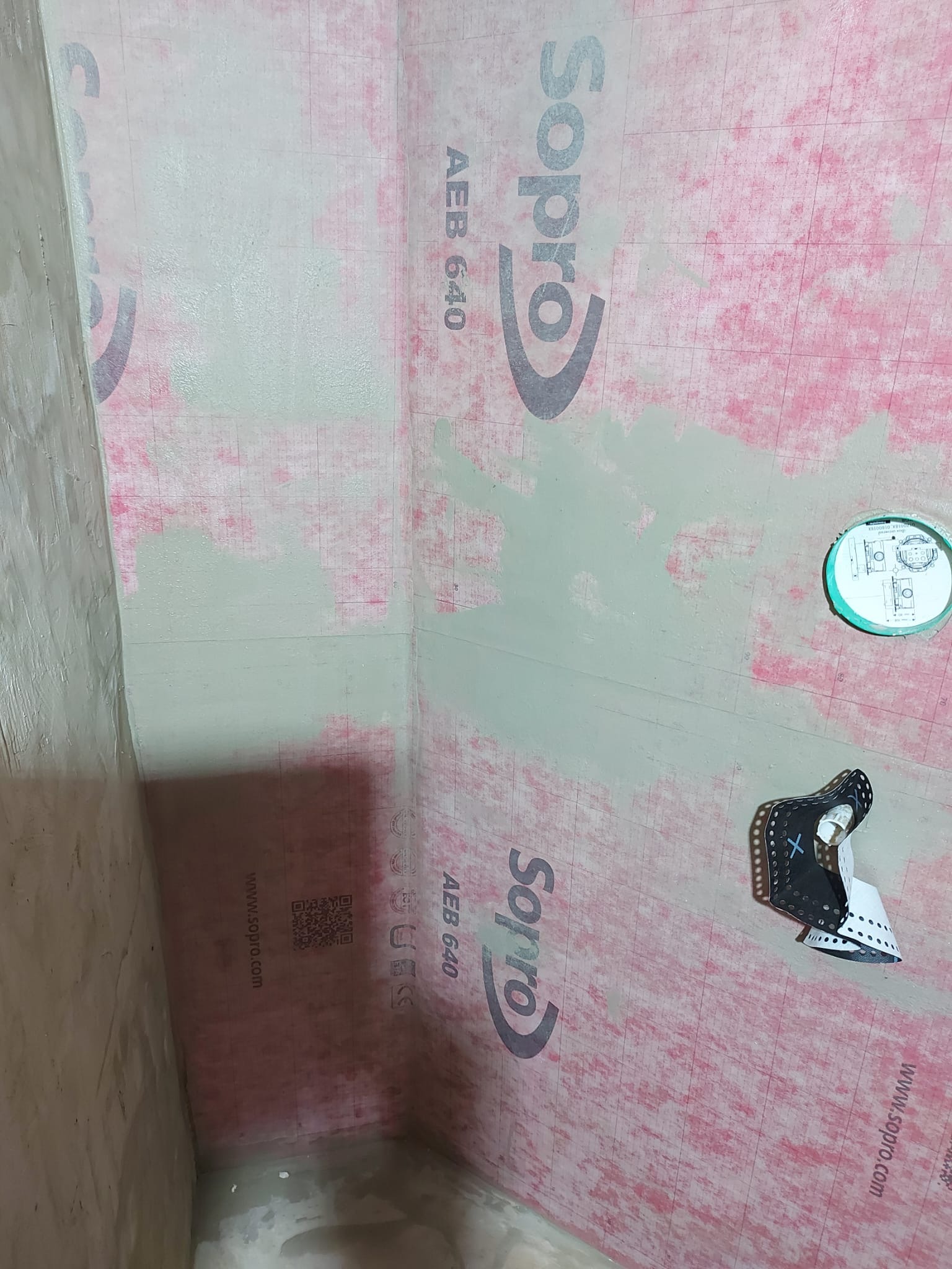 Przygotowanie ścian do prac wykończeniowych w łazience: hydroizolacja Sopro AEB 640 nałożona na ściany, widoczne łączenia zabezpieczone taśmą, odpływ liniowy w podłodze zakryty folią zabezpieczającą.