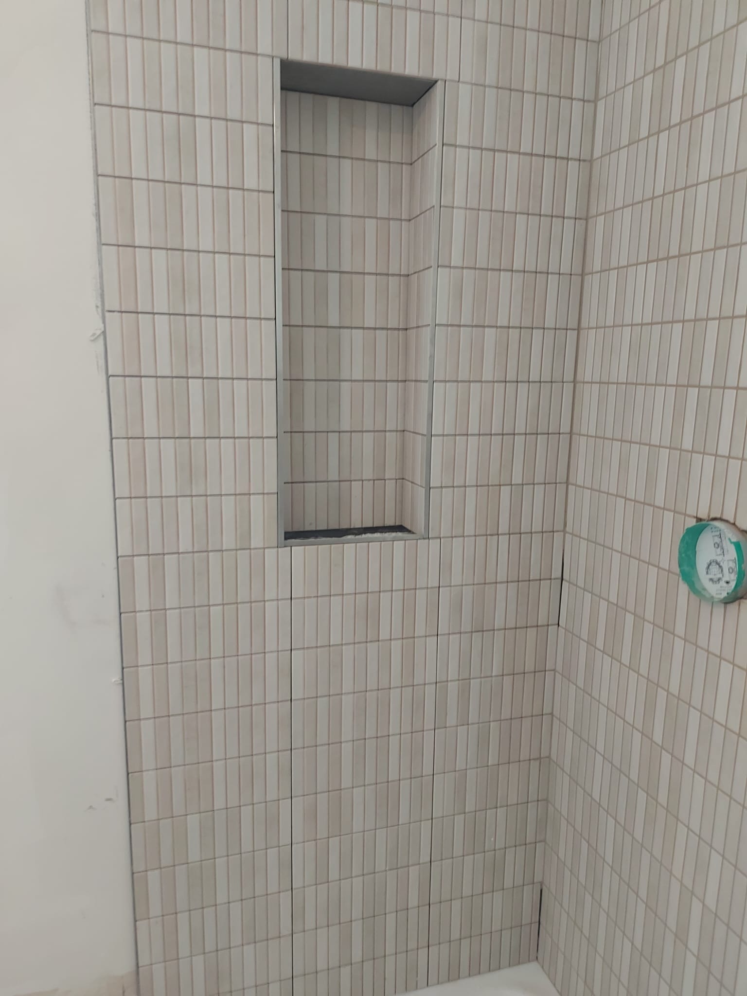 Ściana łazienki wyłożona wąskimi, pionowymi płytkami w odcieniach beżu z wnęką w zabudowie, częściowo wykończona, widoczny niezabudowany fragment ściany oraz osłonięte przyłącze wodne.