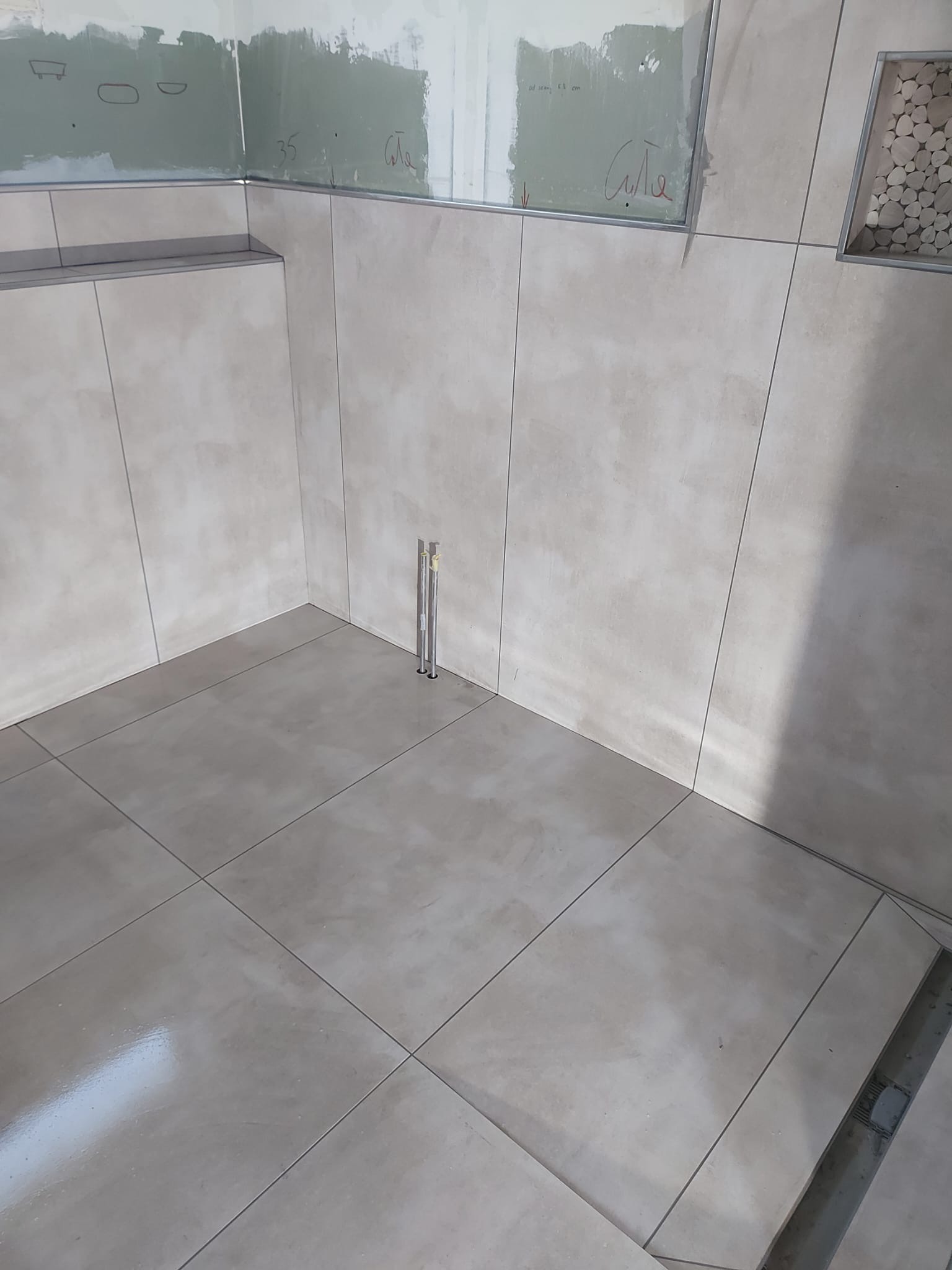 Świeżo wyłożona, beżowa podłoga i ściany z płytek w łazience, widoczne pionowe rury instalacji wodnej i fragment niezabezpieczonej ściany z płyty gipsowo-kartonowej.