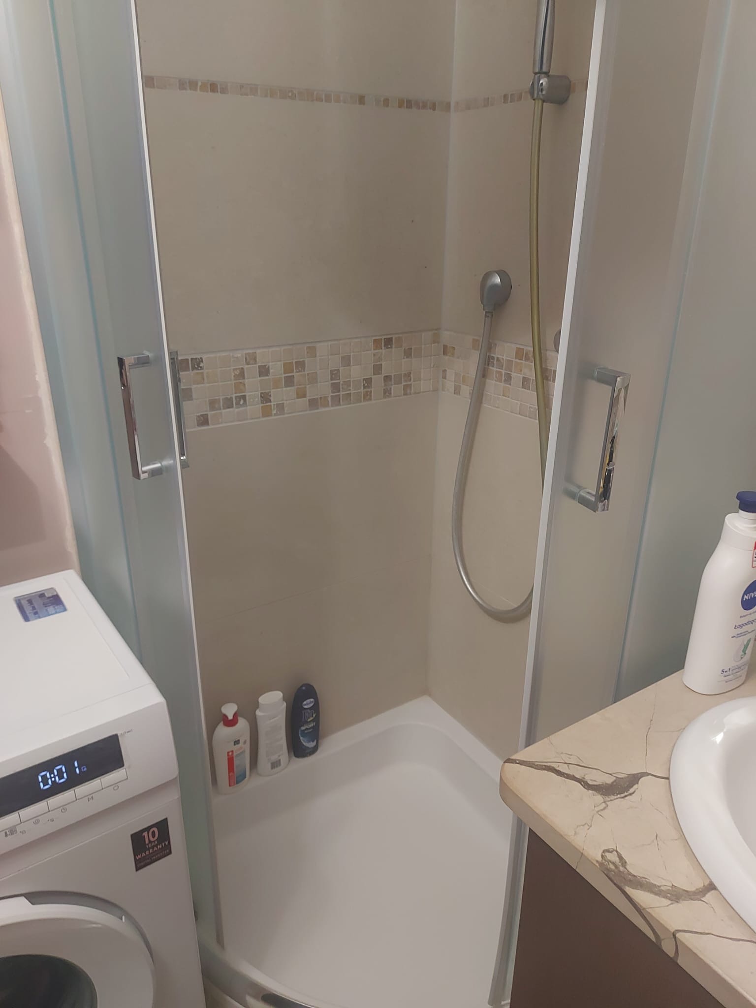 Kabina prysznicowa z przesuwnymi drzwiami, wykończona mozaiką, obok pralka i umywalka z blatem imitującym marmur.