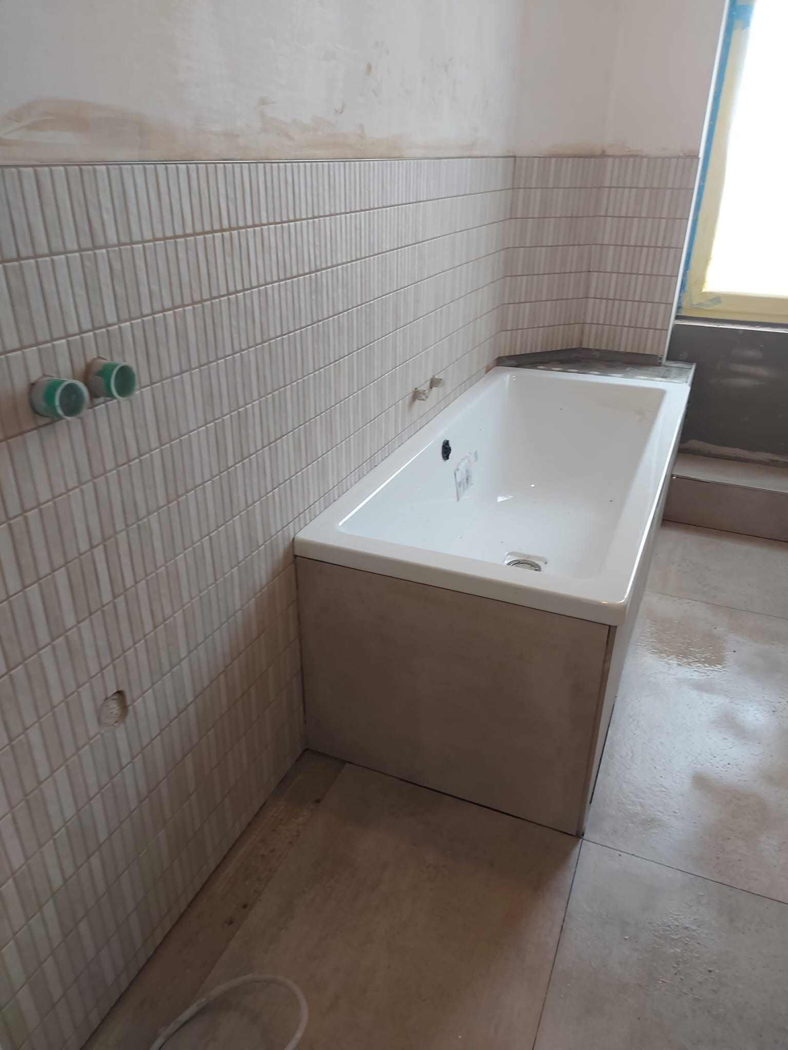 Wanna obudowana płytkami w trakcie montażu, widoczne fragmenty ściany z ułożonymi beżowymi płytkami w pionowe paski, przygotowane przyłącza wodne z zielonymi zaślepkami, niedokończone fugowanie.