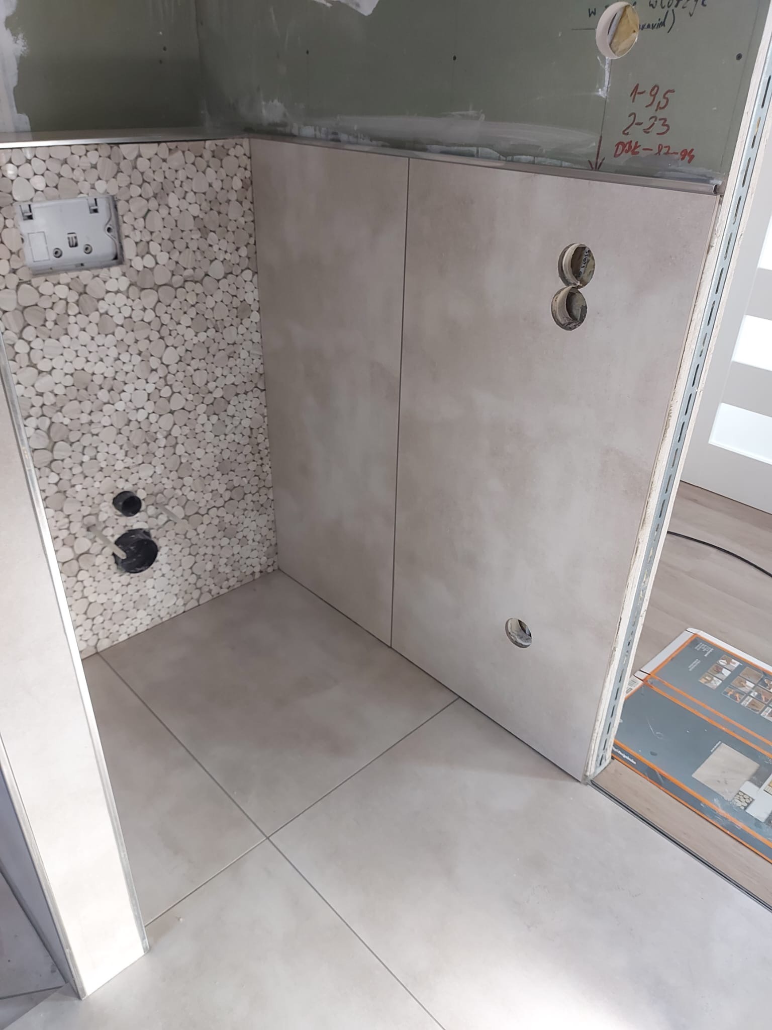 Wykończona ściana łazienki z mozaiką z drobnych kamyczków i gładkimi płytkami, widoczne otwory na instalacje, przygotowana do montażu armatury.