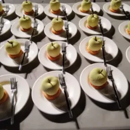 Rząd białych talerzy z deserami w kształcie zielonych jabłek na pomarańczowej podstawie, każdy z widelcem, ustawionych na szarym stole pod ciemnym oświetleniem.