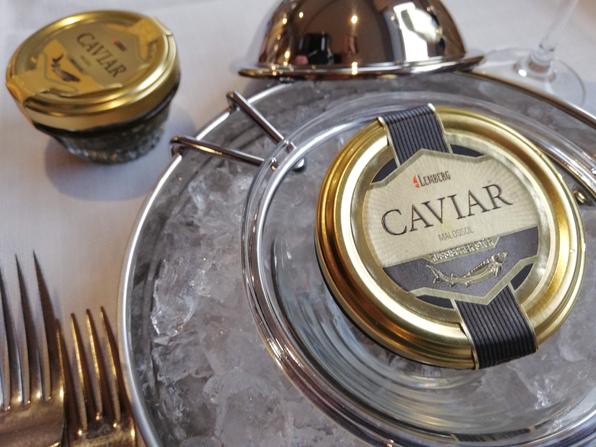 Słoiki z kawiorem na lodzie, widoczne złote wieczka z etykietami marki Lemberg, srebrne sztućce i naczynia w tle.