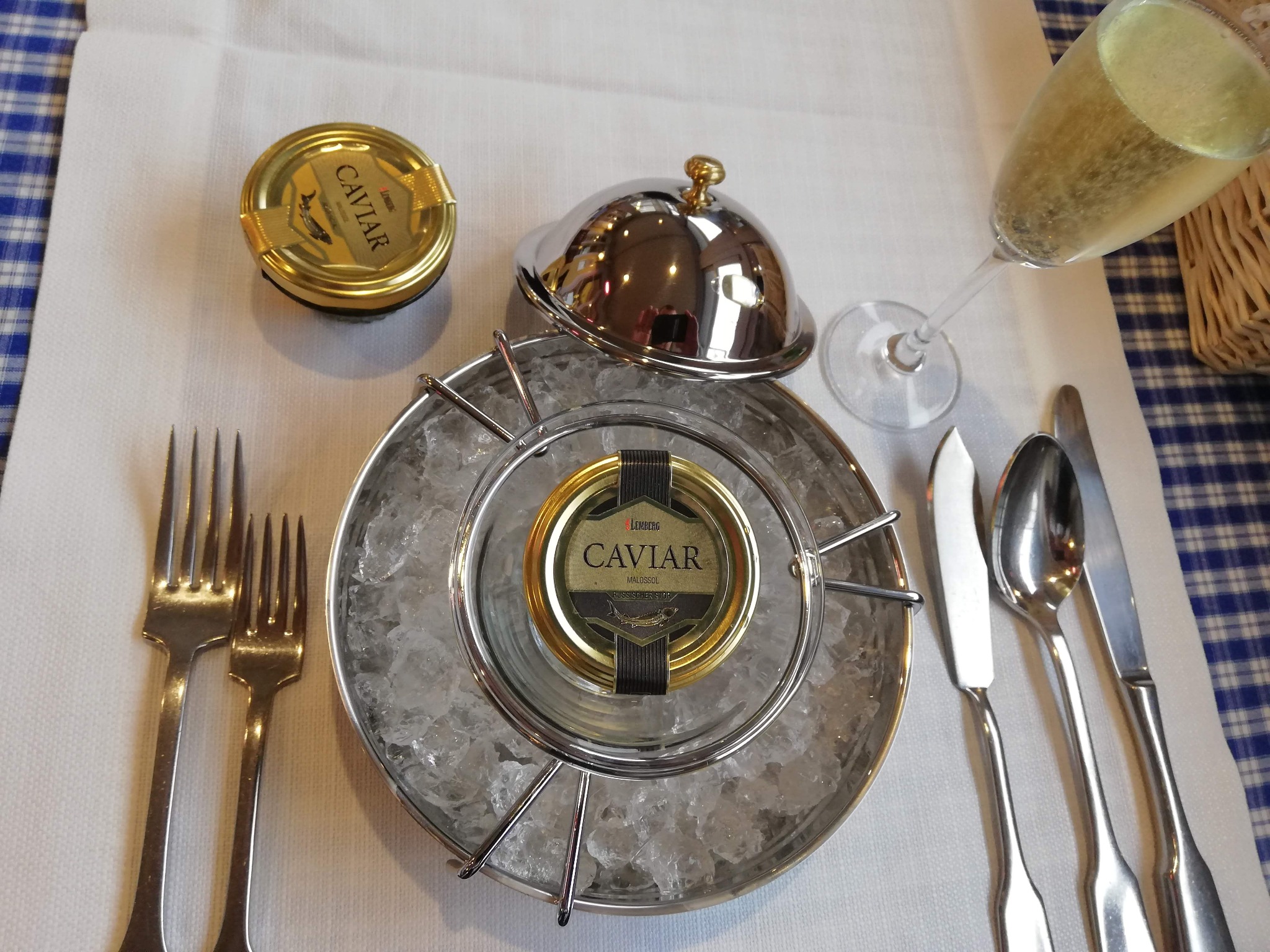 Elegancko podany kawior w metalowej misie z lodem, obok kieliszek szampana, złota puszka kawioru, srebrne sztućce i biały obrus z niebieską kratą.