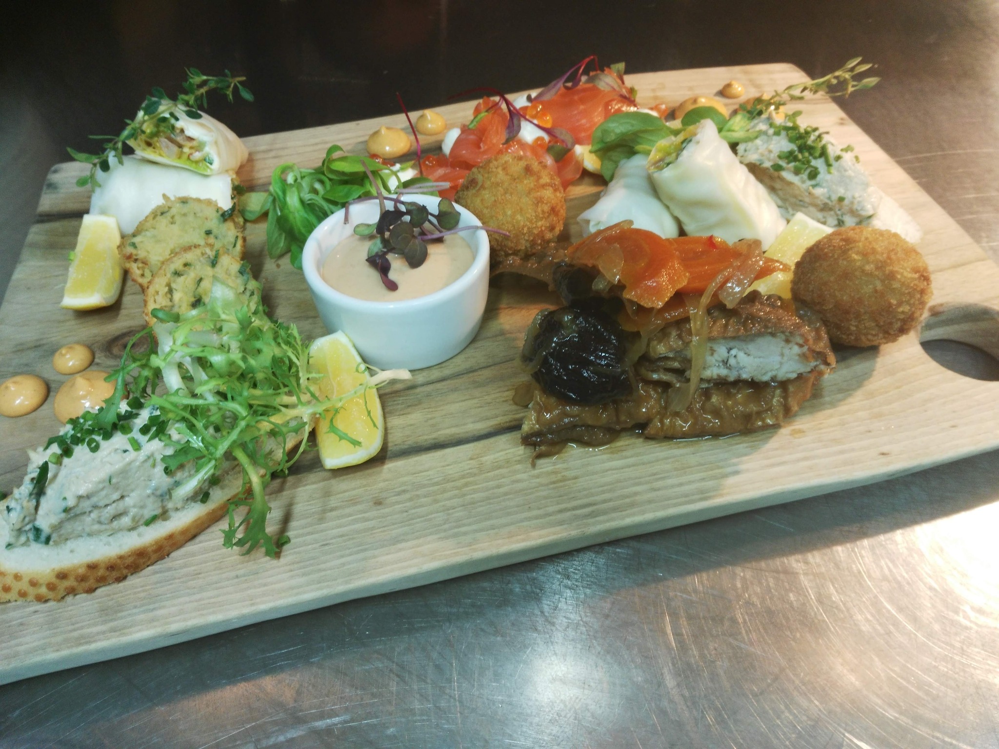 Drewniana deska z różnorodnymi przekąskami: mini tortille, pasta na pieczywie, smażone kulki, łosoś, sos w miseczce, marynowane śliwki i cytryna, udekorowane zieleniną.