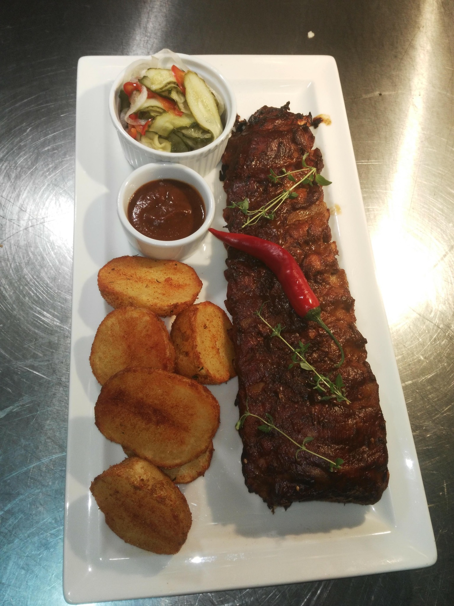 Żeberka BBQ z czerwoną papryczką chili, ziemniakami, sosem i piklami na białym, prostokątnym talerzu, prezentowane na metalowym blacie.