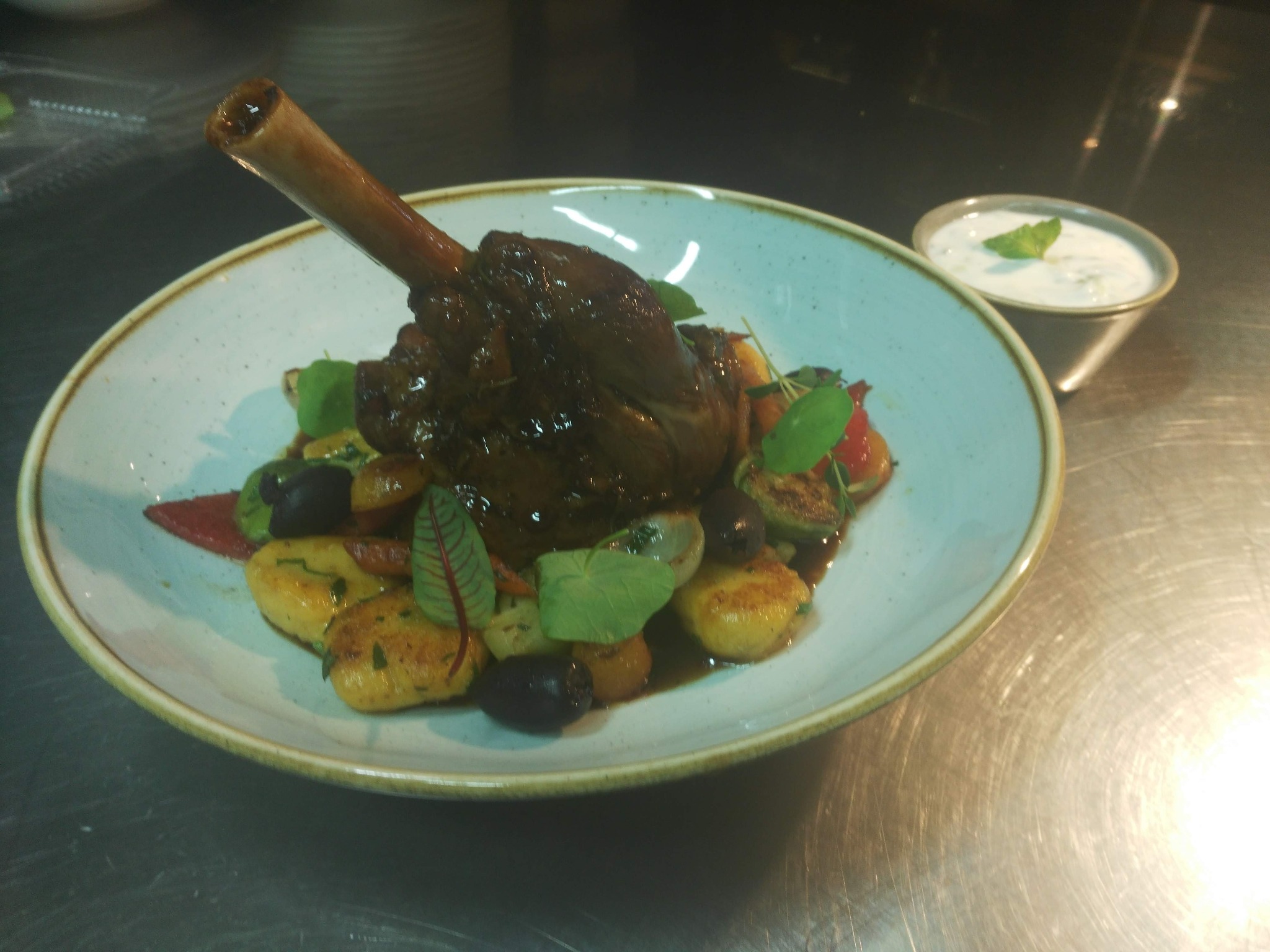 Pieczeń jagnięca z kością na gnocchi z warzywami, podana na jasnym talerzu obok miseczki z sosem miętowym.