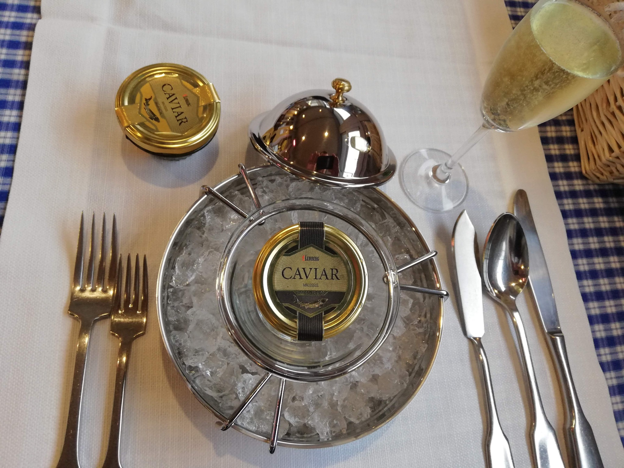 Elegancka prezentacja kawioru Lemberg Malossol w srebrnym naczyniu z lodem, obok kieliszek szampana, srebrne sztućce i biały obrus z niebieską kratą.