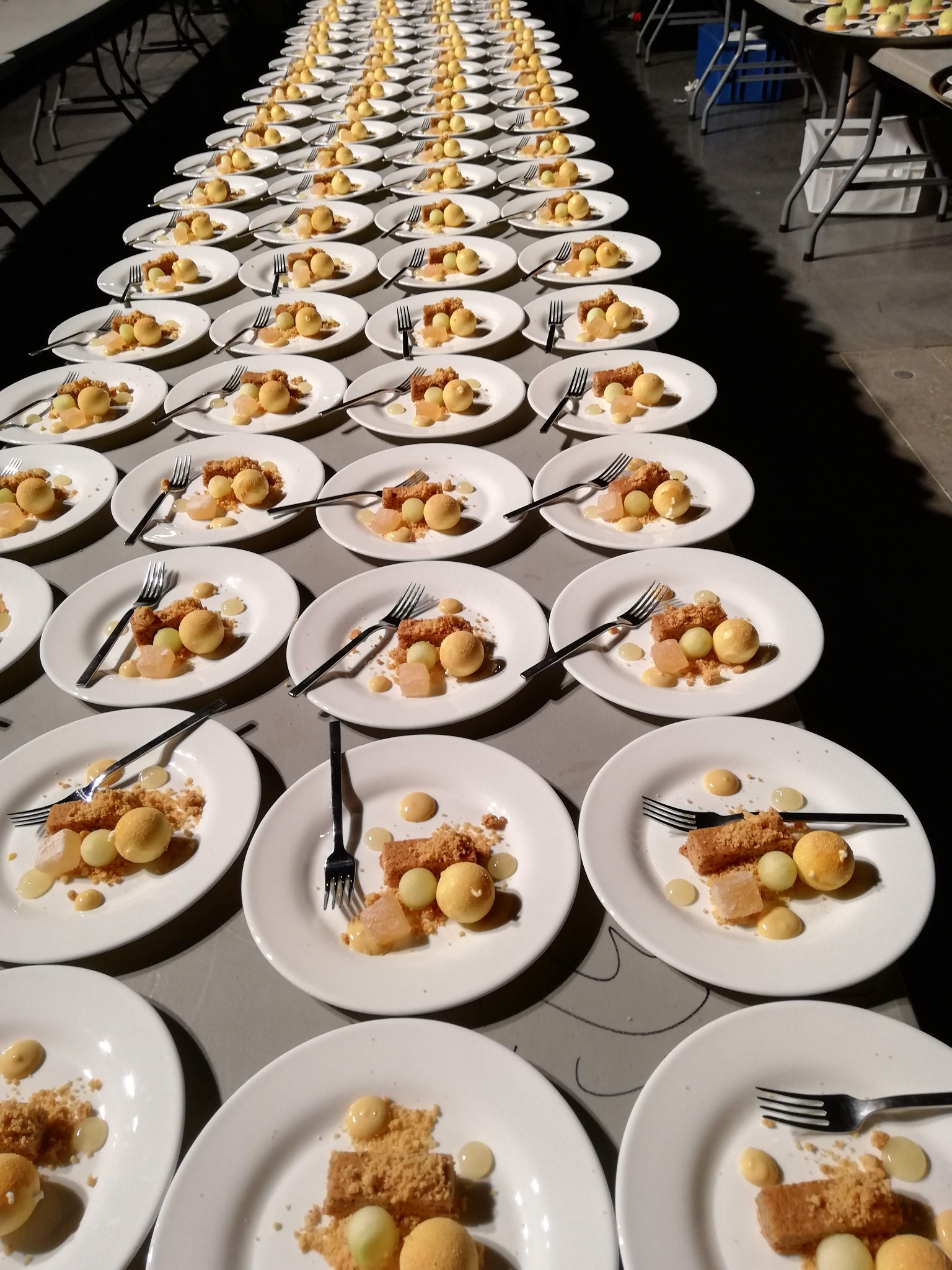 Rząd białych talerzy z deserami: kulki, kostki, kruszonka i sos, każdy talerz z czarnym widelcem, ustawione na szarym stole podczas przygotowań do wydarzenia.