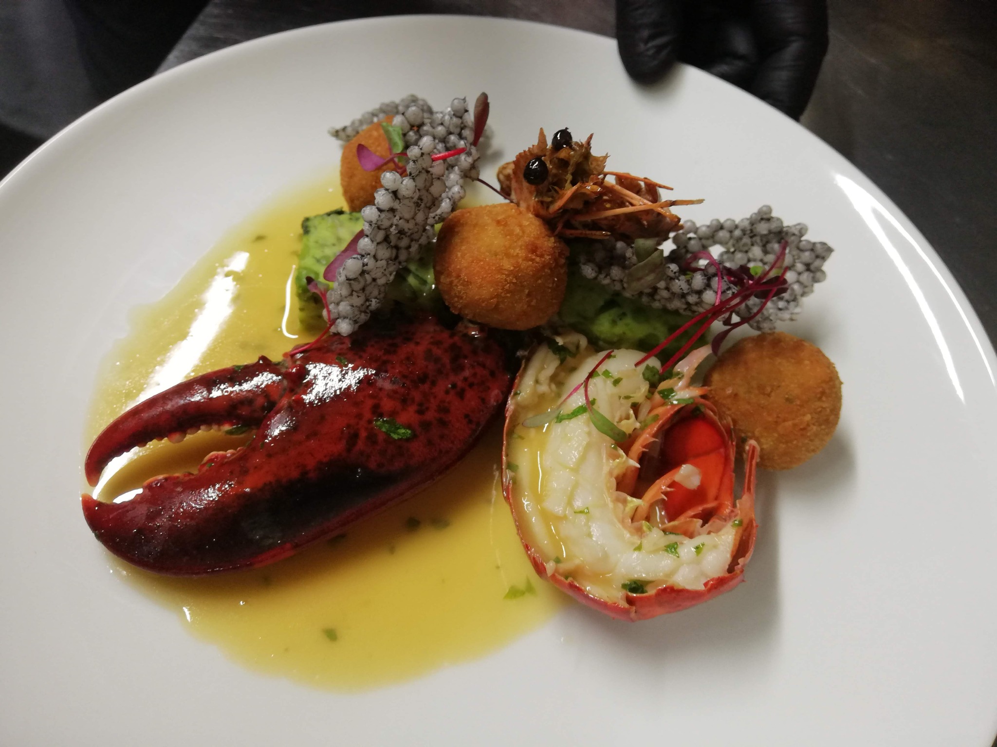 Wykwintne danie z homara: połowa skorupiaka z białym mięsem, szczypce, smażone kulki, zielone puree i dekoracje na białym talerzu, prezentowane przez kucharza w czarnych rękawiczkach.