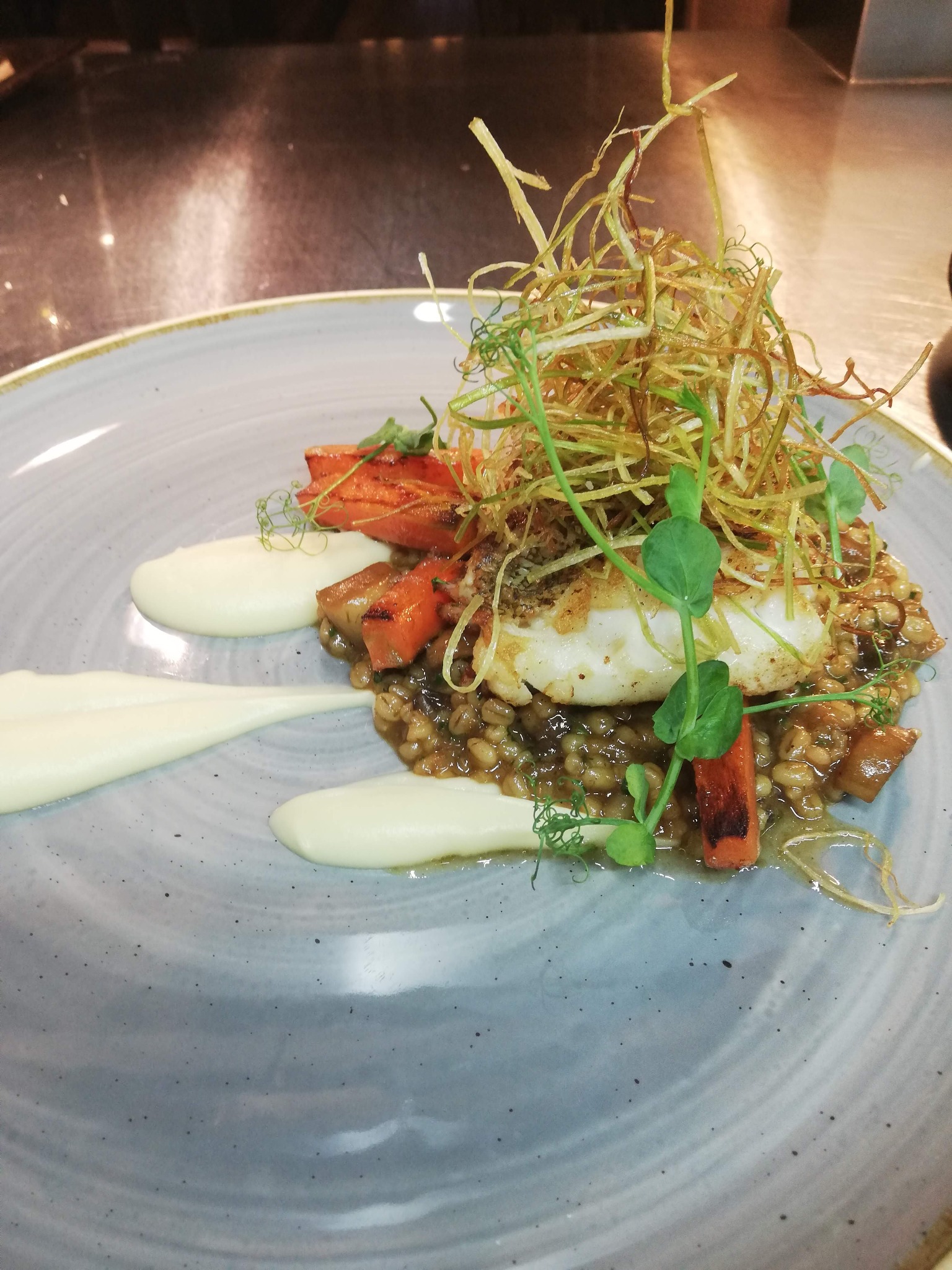 Elegancko podane danie: Filet rybny na kaszy z warzywami, dekorowany sosem, smażonymi porami i świeżymi pędami groszku, na szarym talerzu.