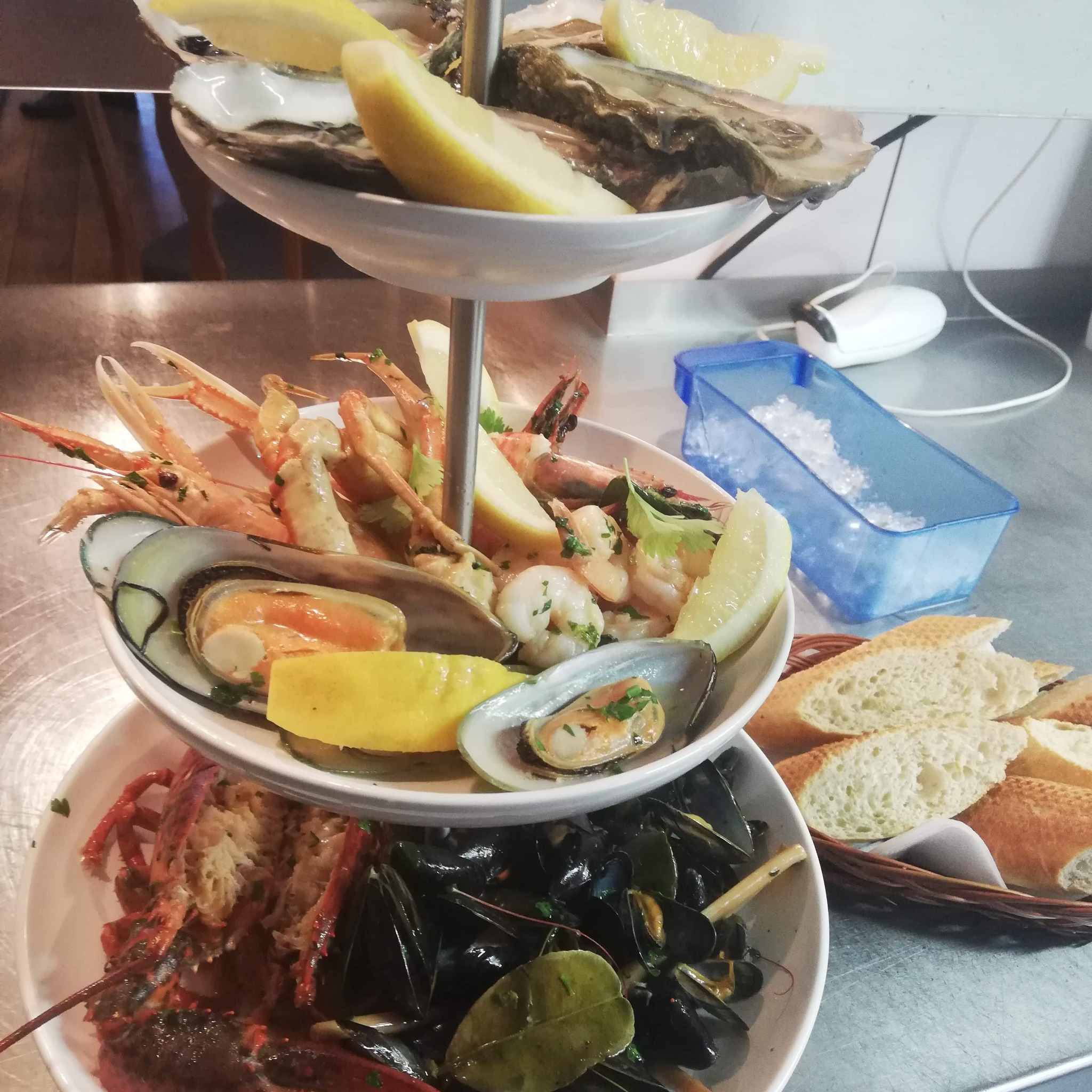 Trzypoziomowy pater z owocami morza: ostrygi z cytryną, krewetki, mule i homar, obok koszyk z bagietką i pojemnik z lodem.