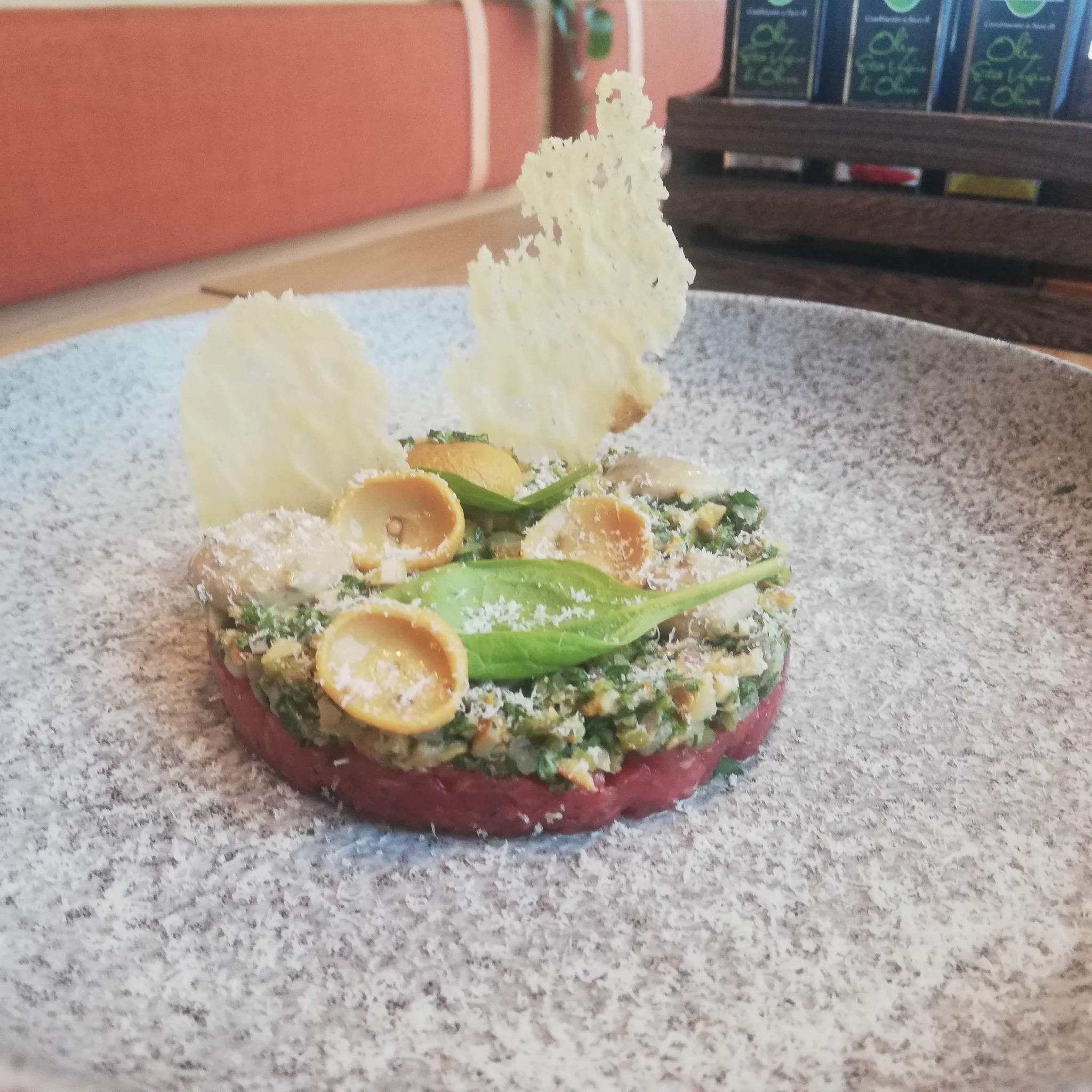 Tatar wołowy z kaparami, ziołami i chipsami parmezanowymi, udekorowany listkami szpinaku i makaronem conchiglie, podany na talerzu posypanym parmezanem, w tle oliwy smakowe.