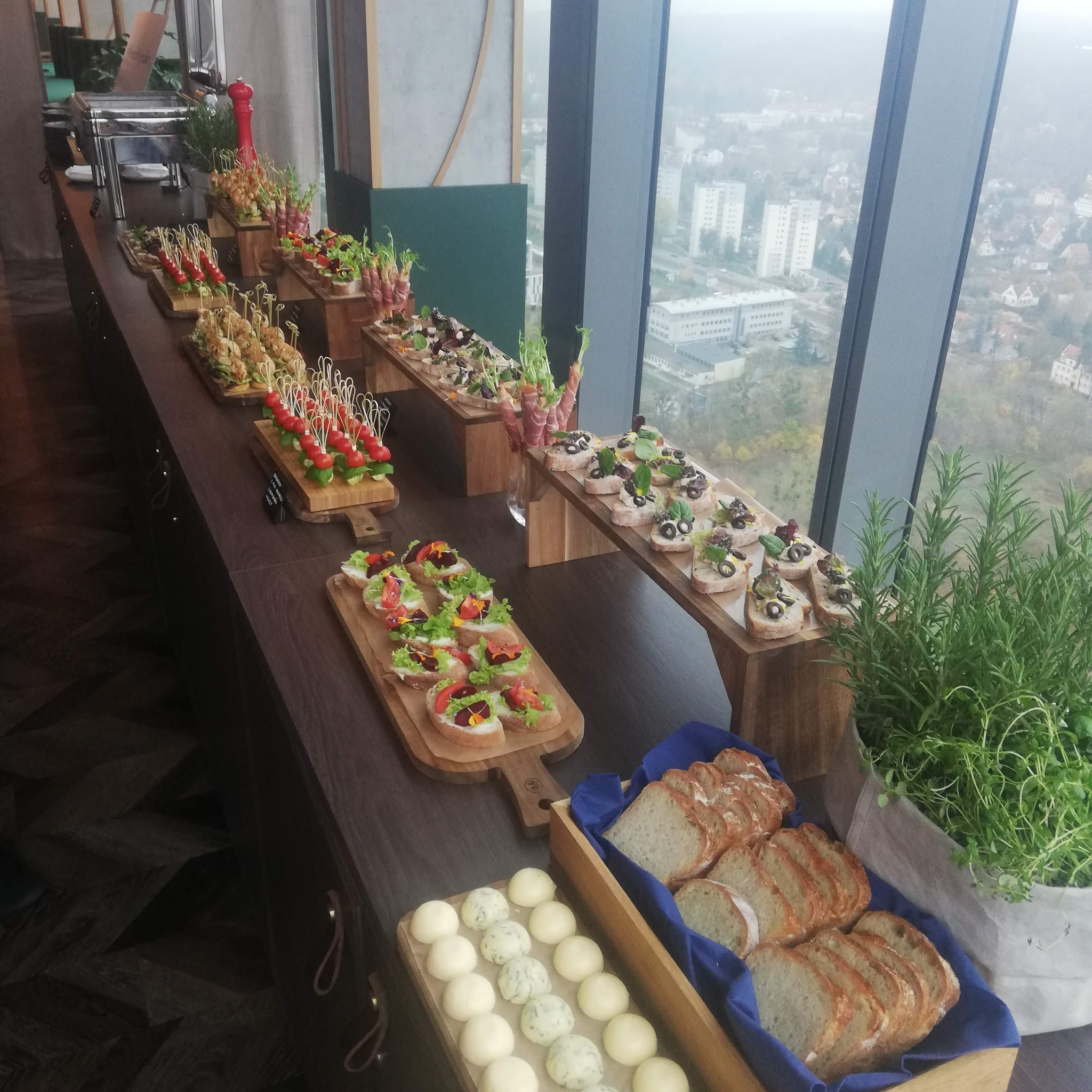 Bufet z różnorodnymi przekąskami na drewnianych deskach i podwyższeniach, widok z okna na miasto w tle, świeże zioła w doniczce obok pieczywa.