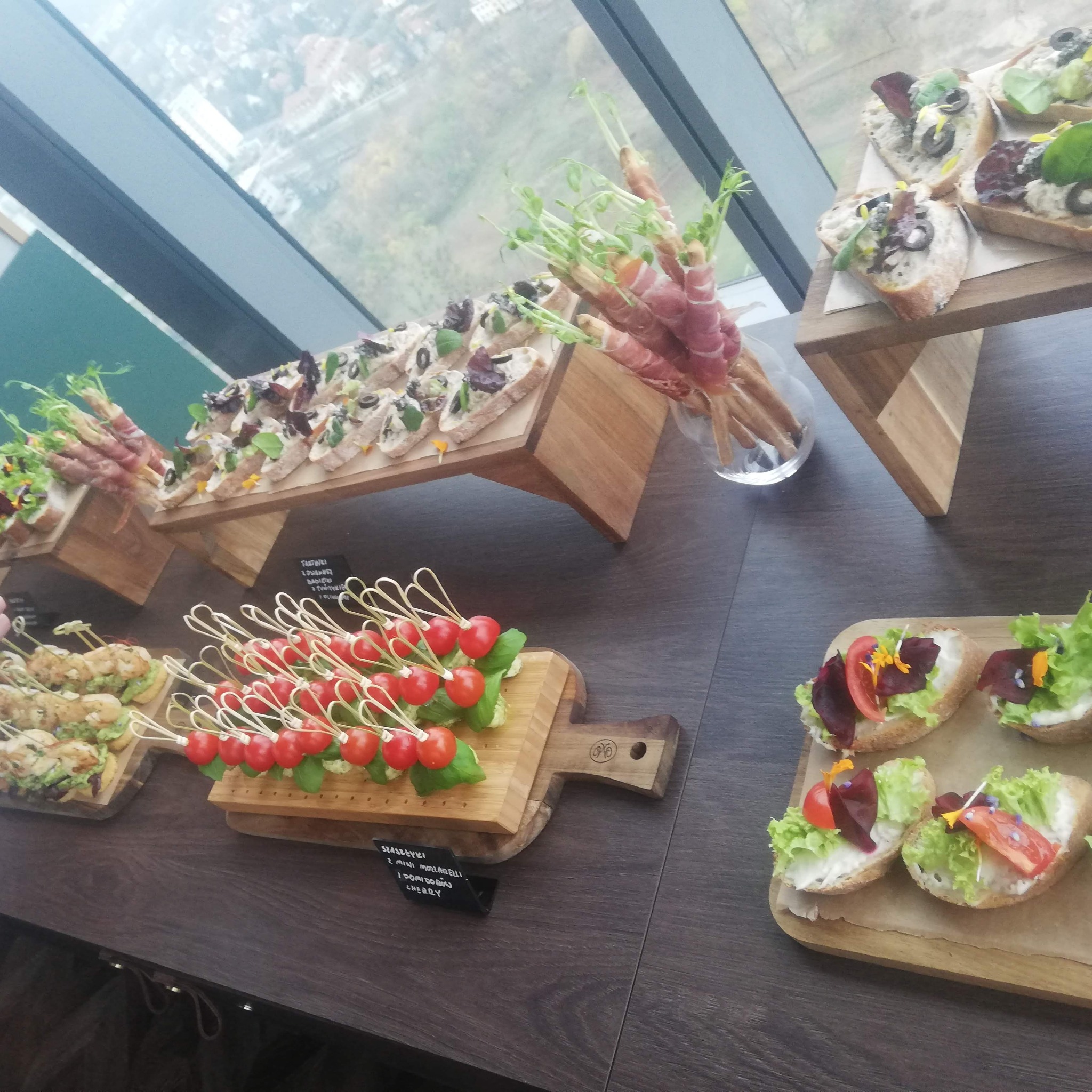 Różnorodne przekąski na drewnianych deskach i podstawkach, w tle widok z okna na miasto.