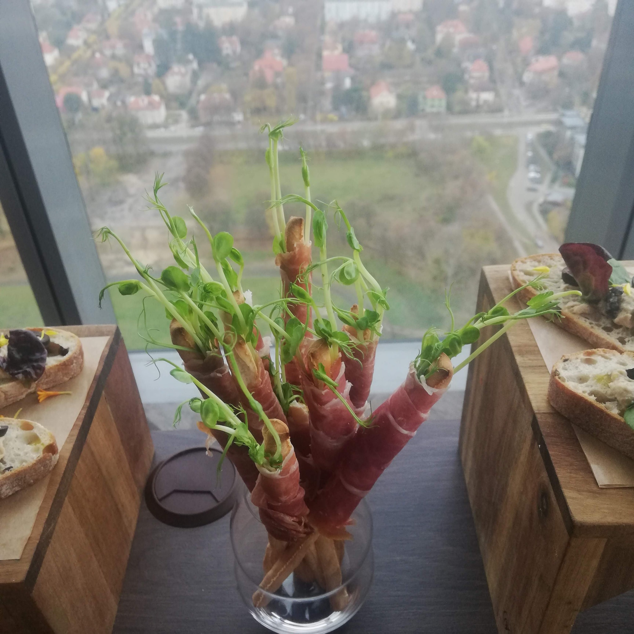 Elegancka kompozycja przekąsek na przyjęciu: szynka parmeńska owinięta wokół paluszków grissini, udekorowana świeżymi pędami groszku, podana w szklanym naczyniu na tle widoku miasta z wysokości.