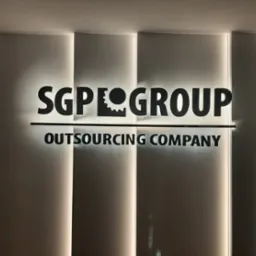 Świecący napis SGP GROUP Outsourcing Company na ścianie, podświetlony od tyłu, obok zamontowane elementy metalowe, pionowe linie podświetlenia po bokach.