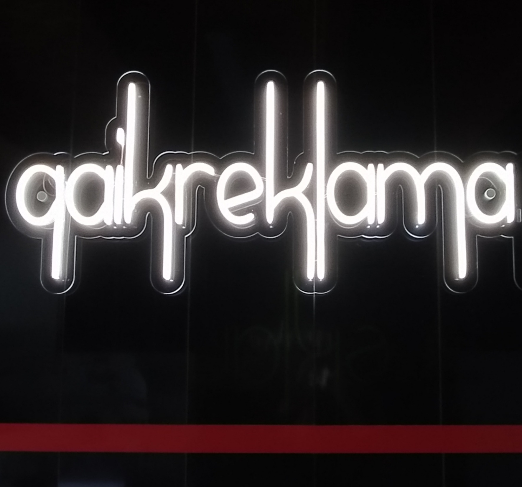 Neonowy napis 'qakreklama' na czarnym tle z czerwoną linią na dole, wykonany w technologii FLEX.